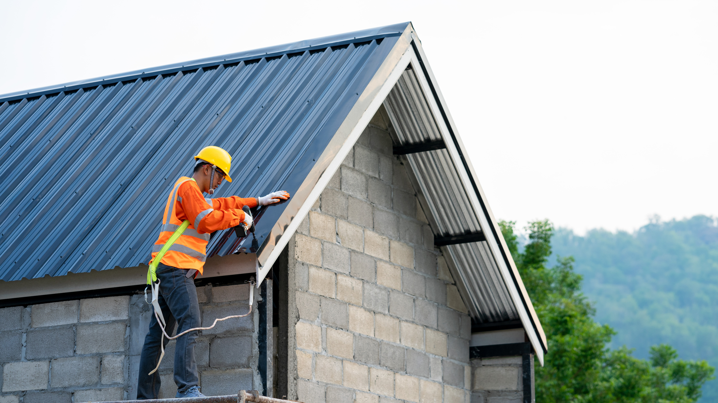 A construction worker wearing an orange safety vest, yellow helmet, and gloves is installing or inspecting corrugated metal roofing on a concrete block building, with green trees and distant hills in the background.