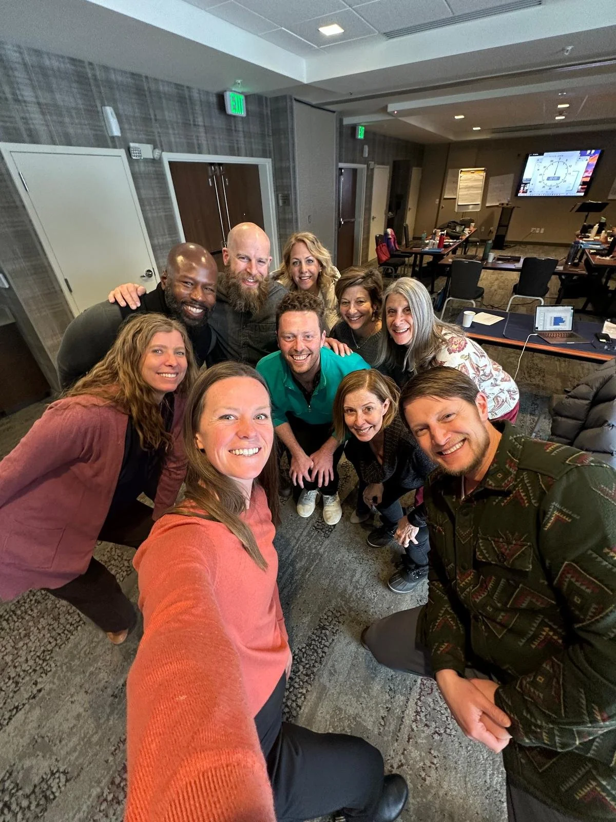 Nina Cashman leading a leadership training workshop with a group of ten happy adults in a conference room taking a group selfie, with tables, chairs, and a large screen displaying a presentation in the background.