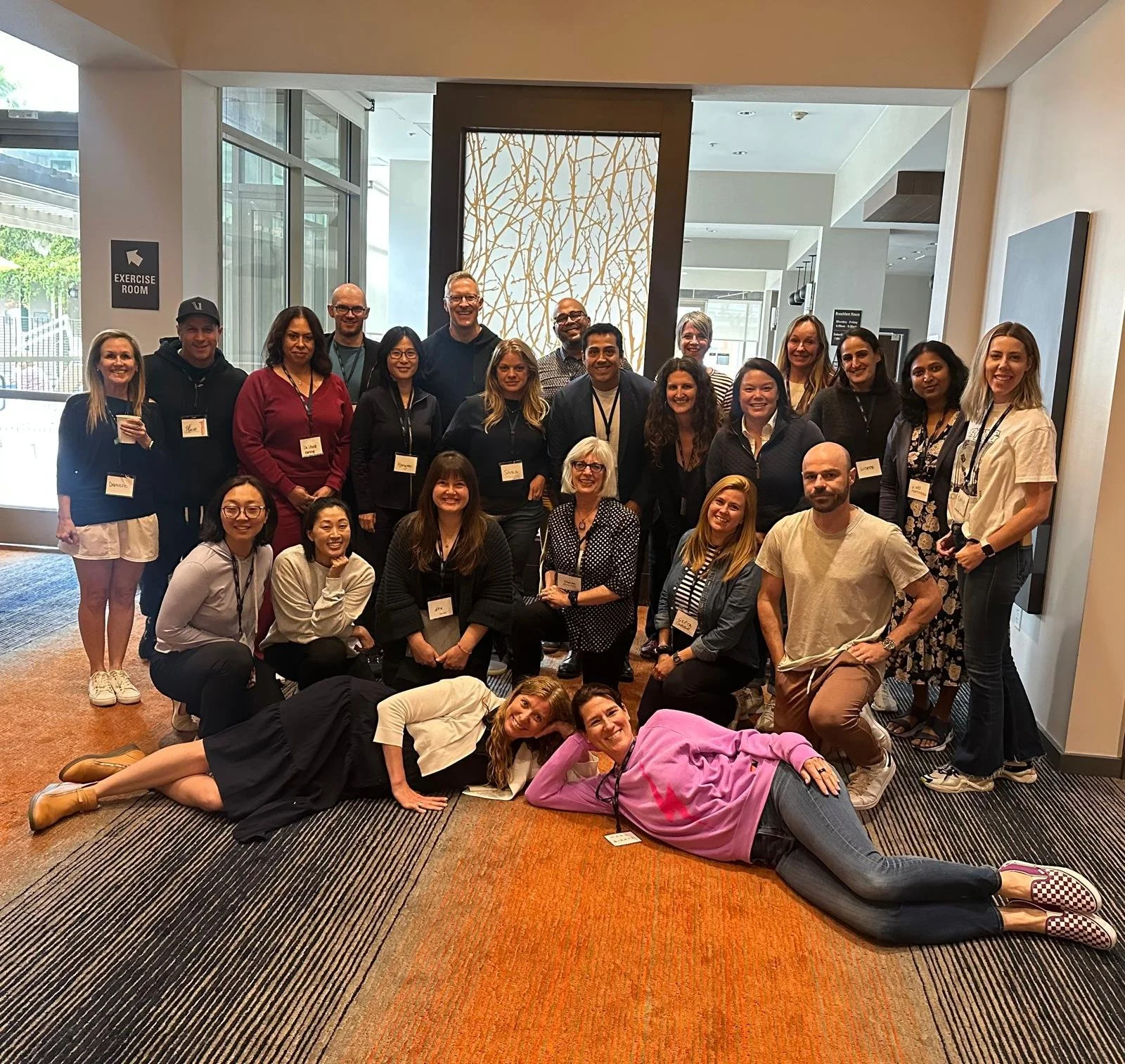 Nina Cashman, certified executive with a group of people gathered for a photo in an indoor setting near a glass entrance. Some are standing, some are sitting or lying on the carpet, all wearing name tags, smiling