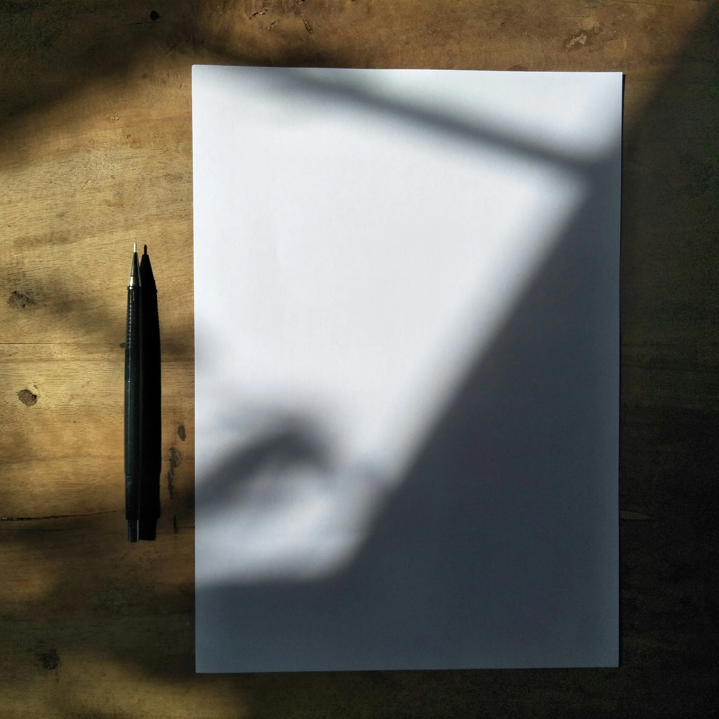 A white sheet of paper on a wooden surface with two black pens placed next to it, partially shadowed by a shadow cast from above.