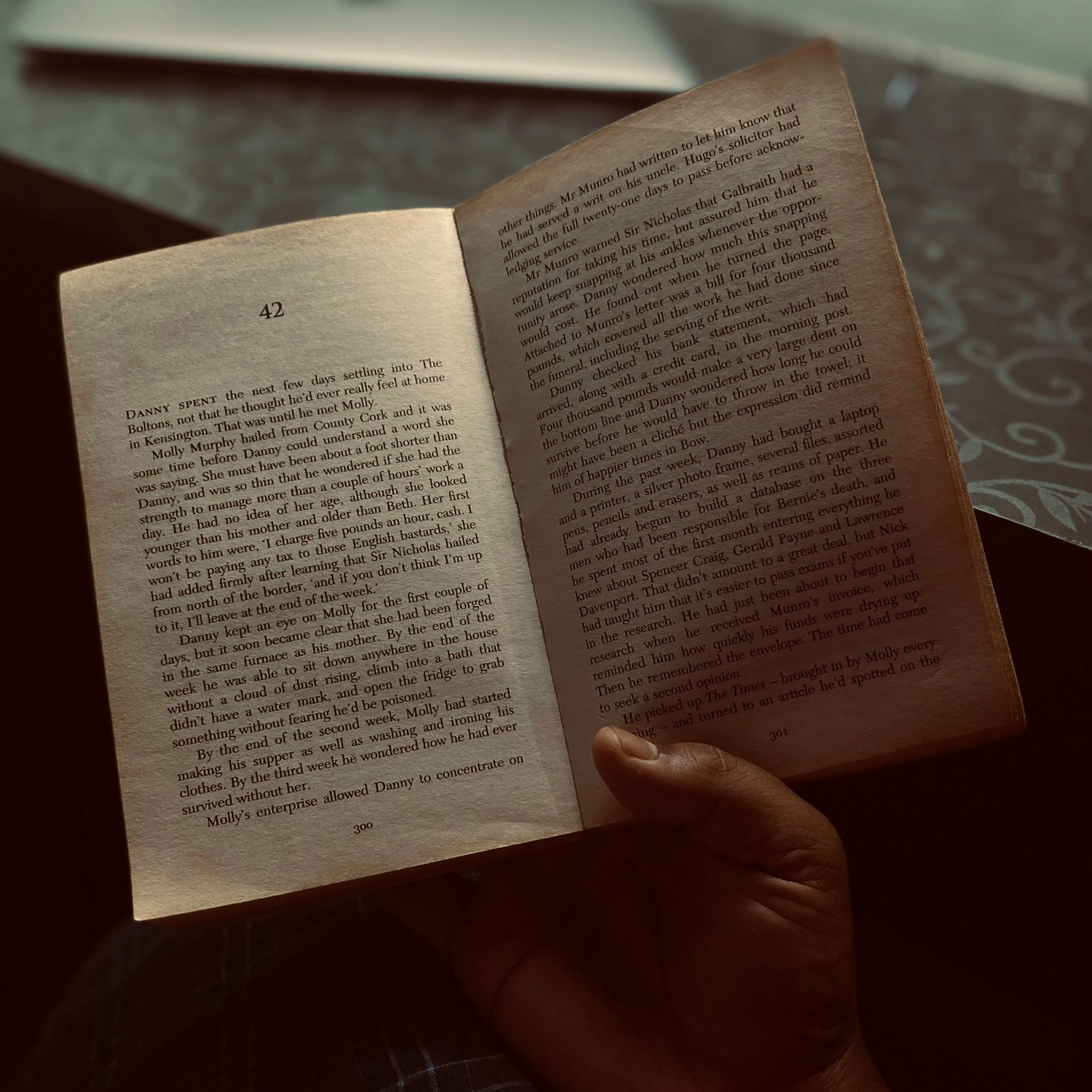 A person holding an open paperback book with pages 300 and 301 visible. The person’s hand is gripping the bottom right corner of page 301. The background shows a table surface with a grey pattern.