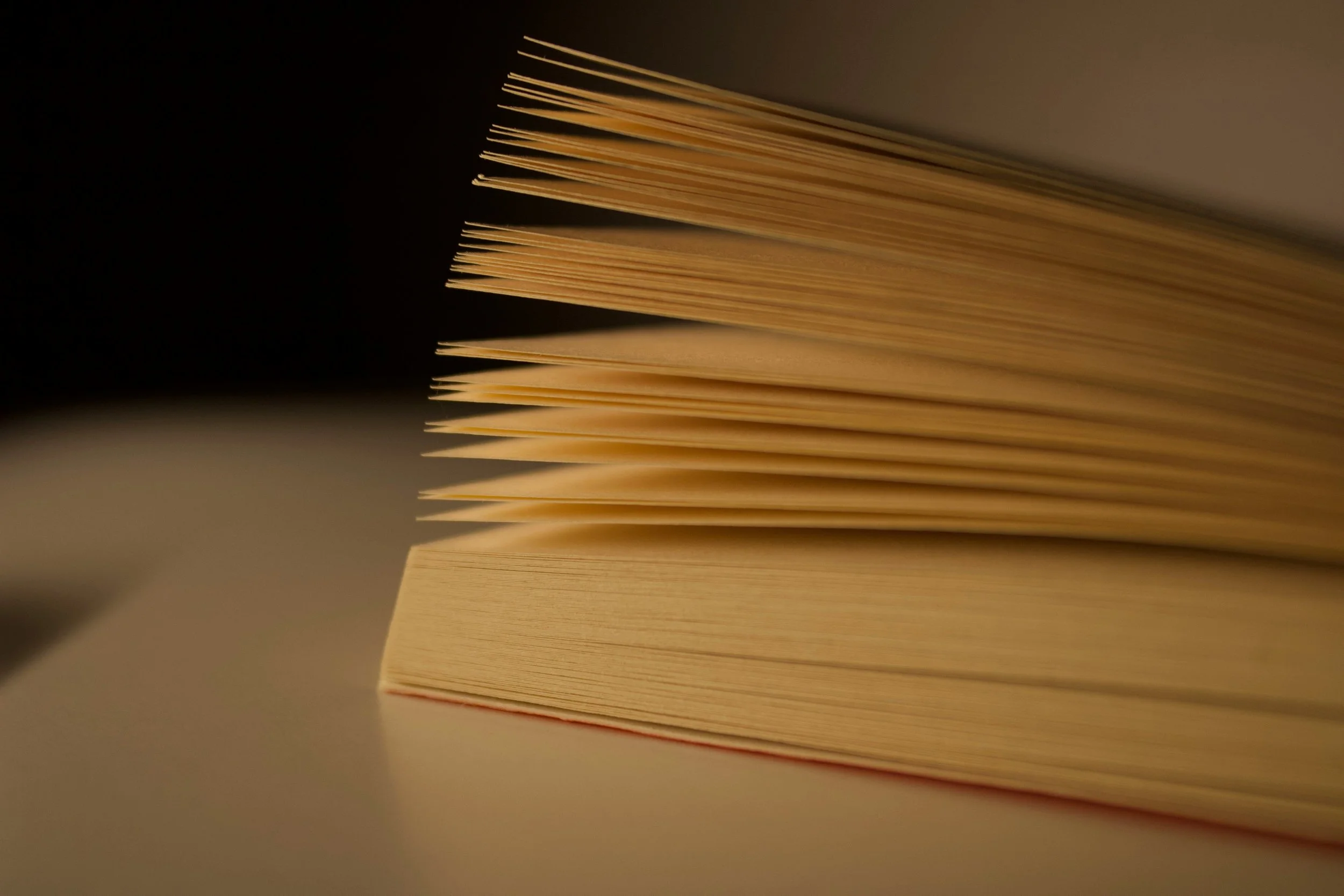Close-up of a stack of cream-colored paper sheets viewed from the side, illuminated with soft lighting, against a dark background.