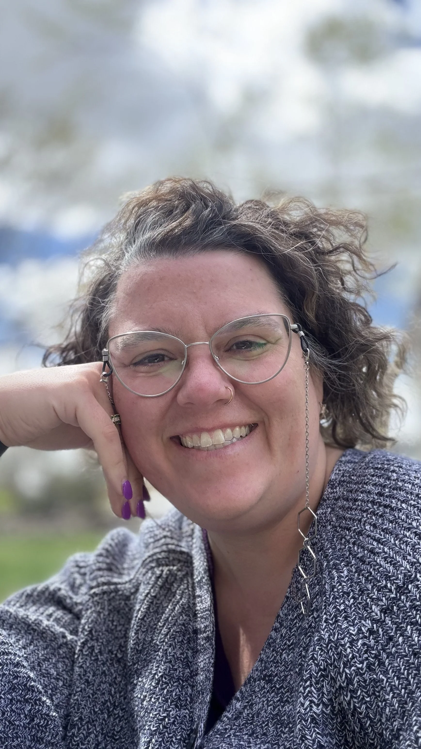 A woman with curly hair, glasses, and a nose ring smiling outdoors. She is wearing a gray textured jacket and has purple nail polish. The background is blurred, showing trees and sky.