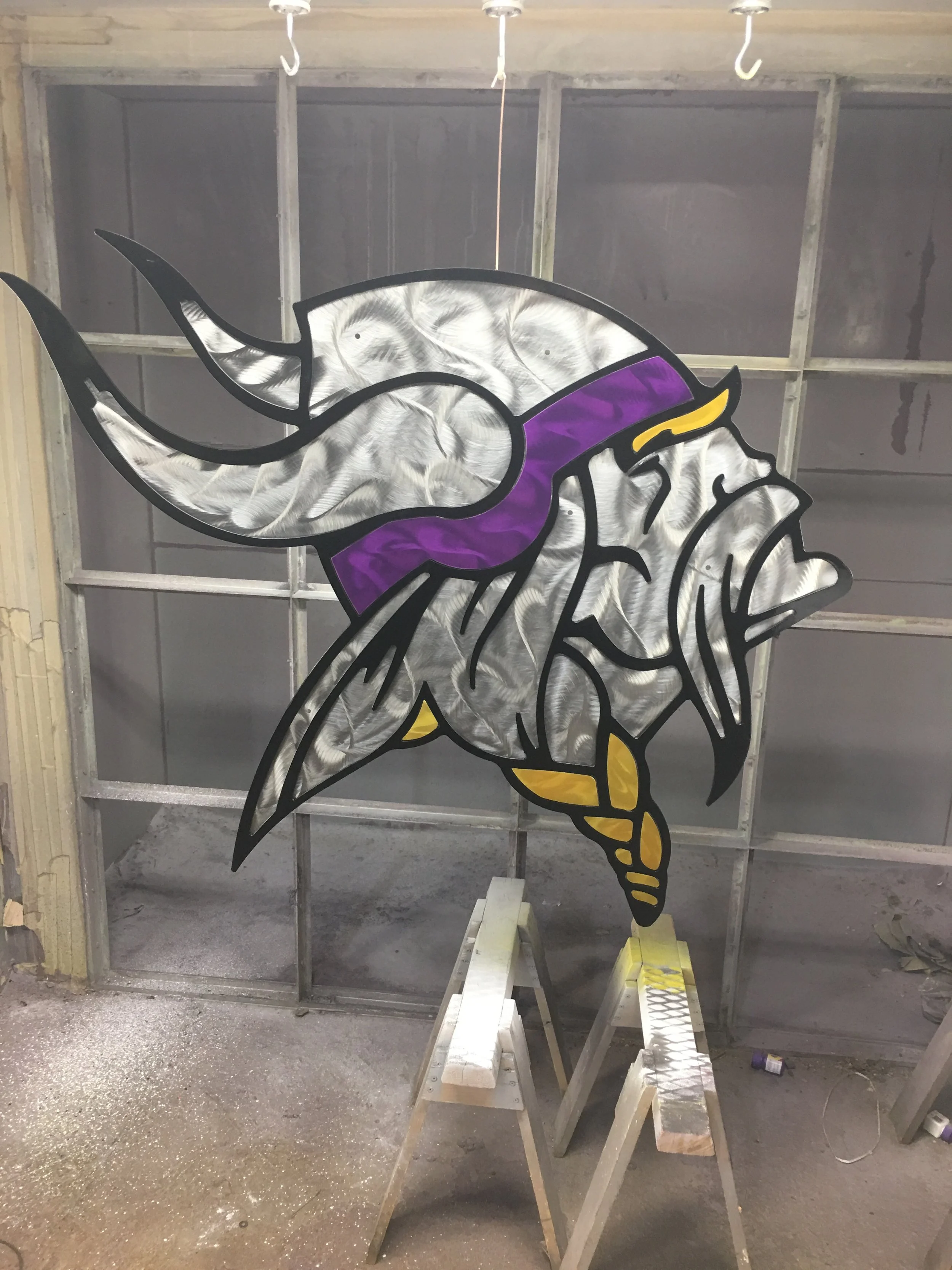 Stained glass window depicting a Viking head with a silver helmet, purple headband, and yellow accents, placed on a workbench in a garage or workshop.