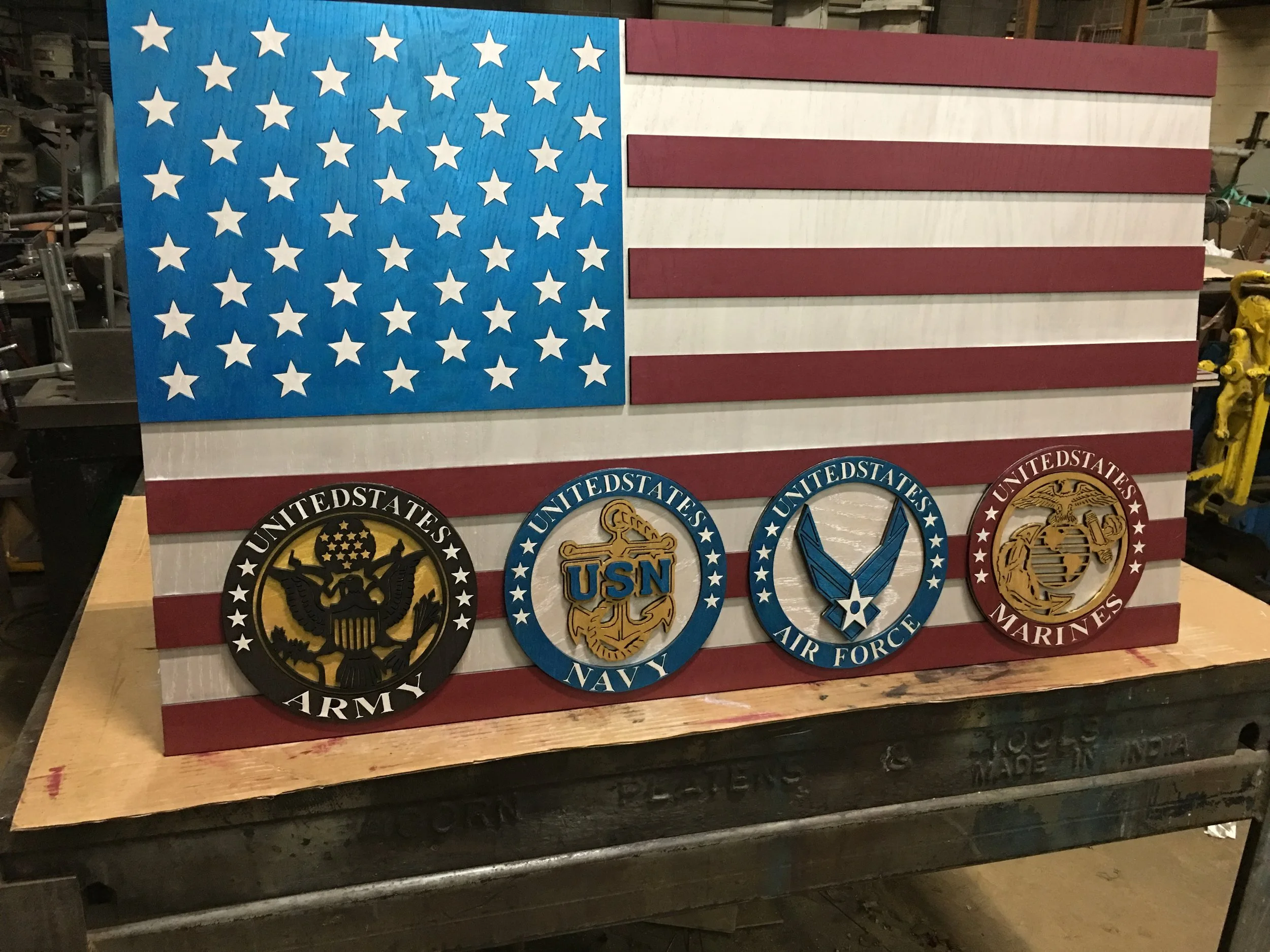 Wooden American flag plaque with seals of the U.S. Army, Navy, Air Force, and Marines displayed on the red and white striped background.