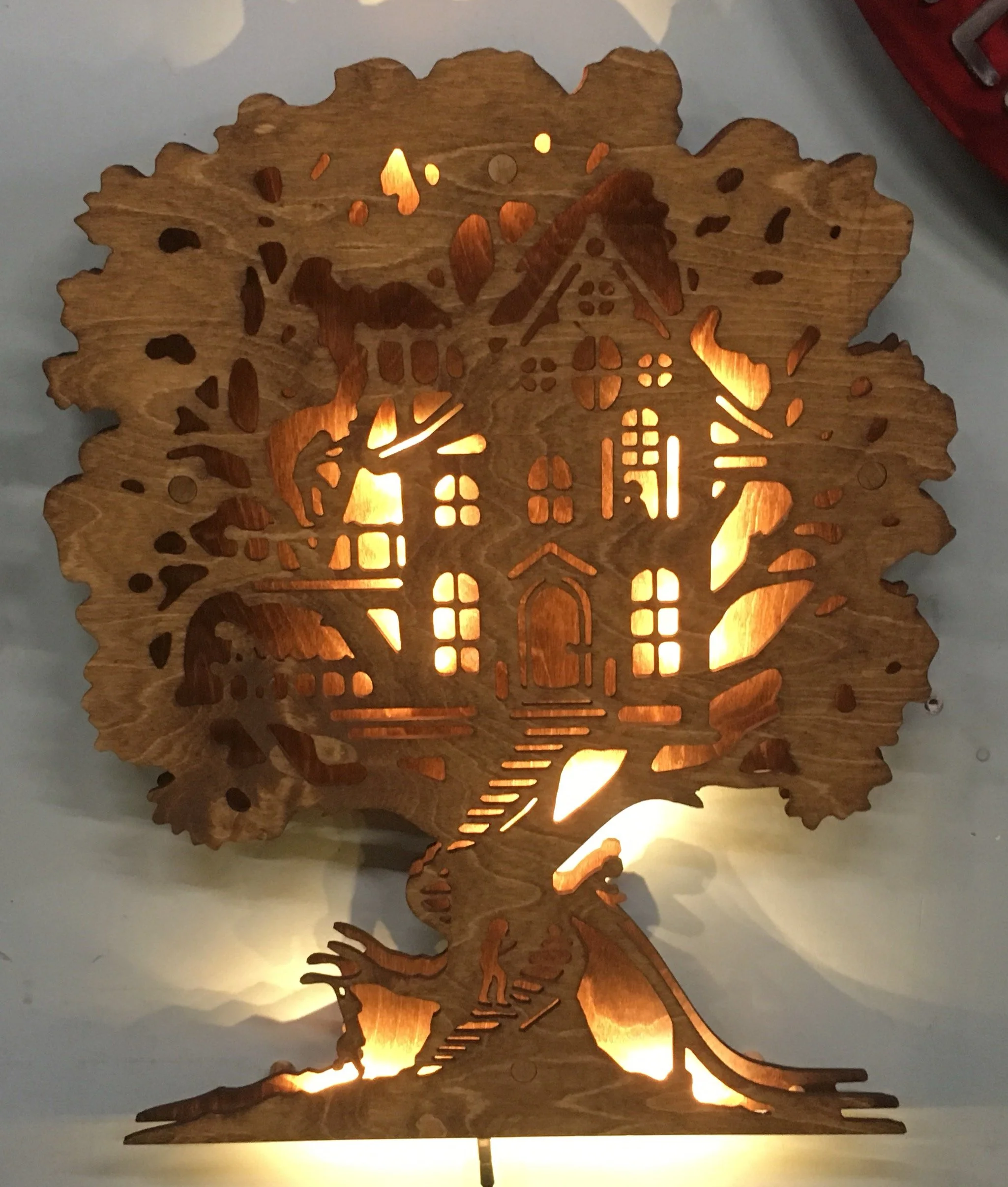 Wooden lamp with cutout design of a house inside a tree, emitting light from within.