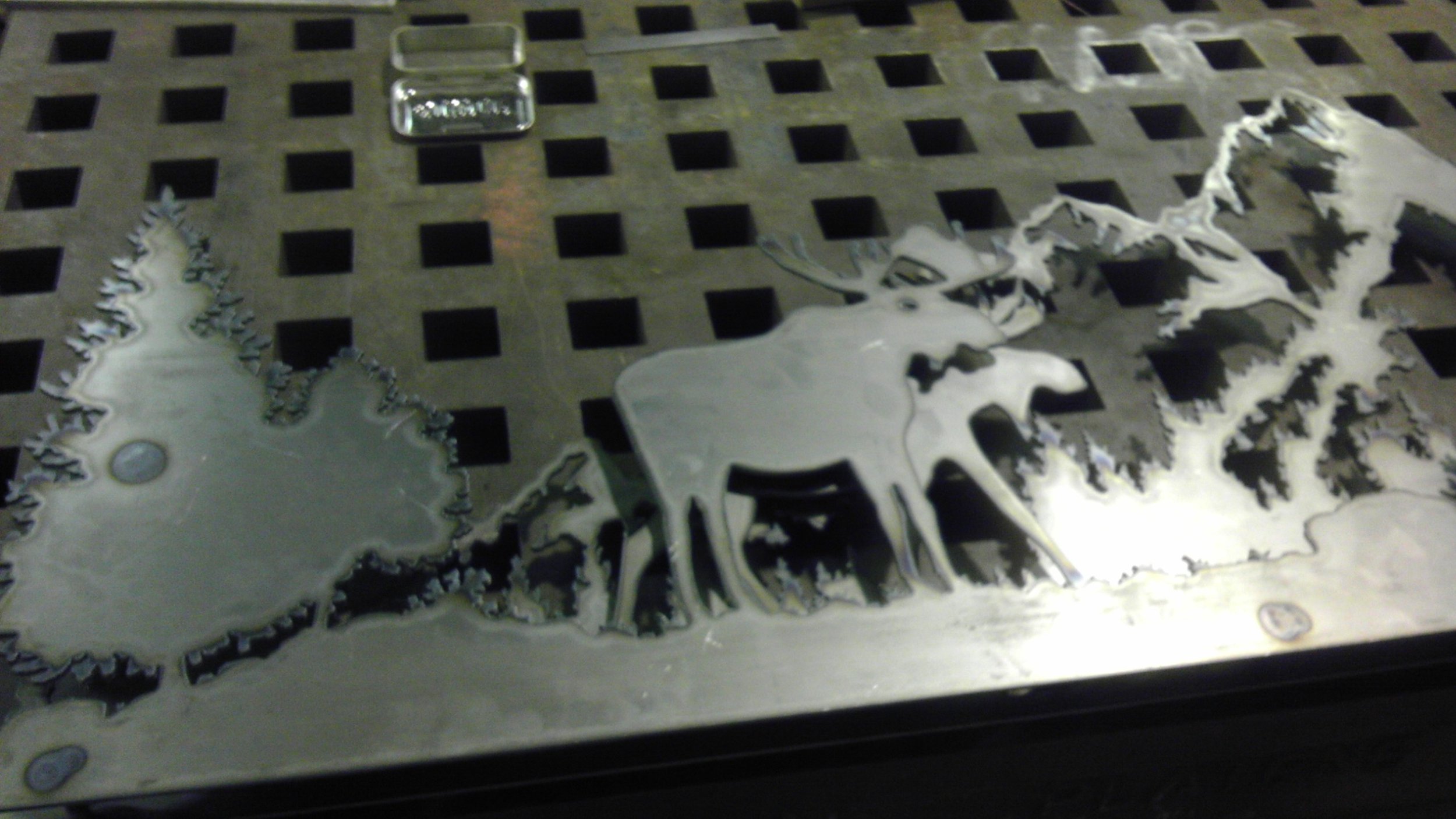 Metal artwork depicting a reindeer and pine trees, placed on a metal grid surface with a small glass container nearby.