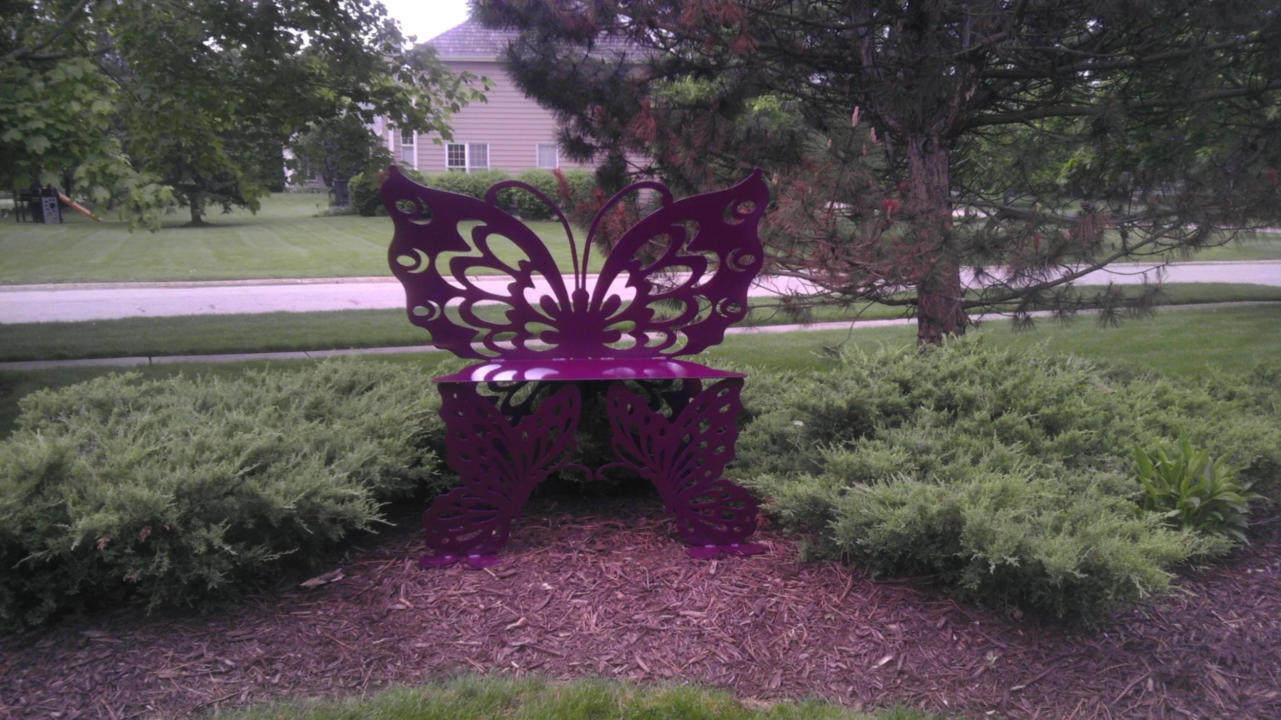 Purple butterfly-shaped outdoor decorative chair situated amidst green bushes with a tree and residential houses in the background.