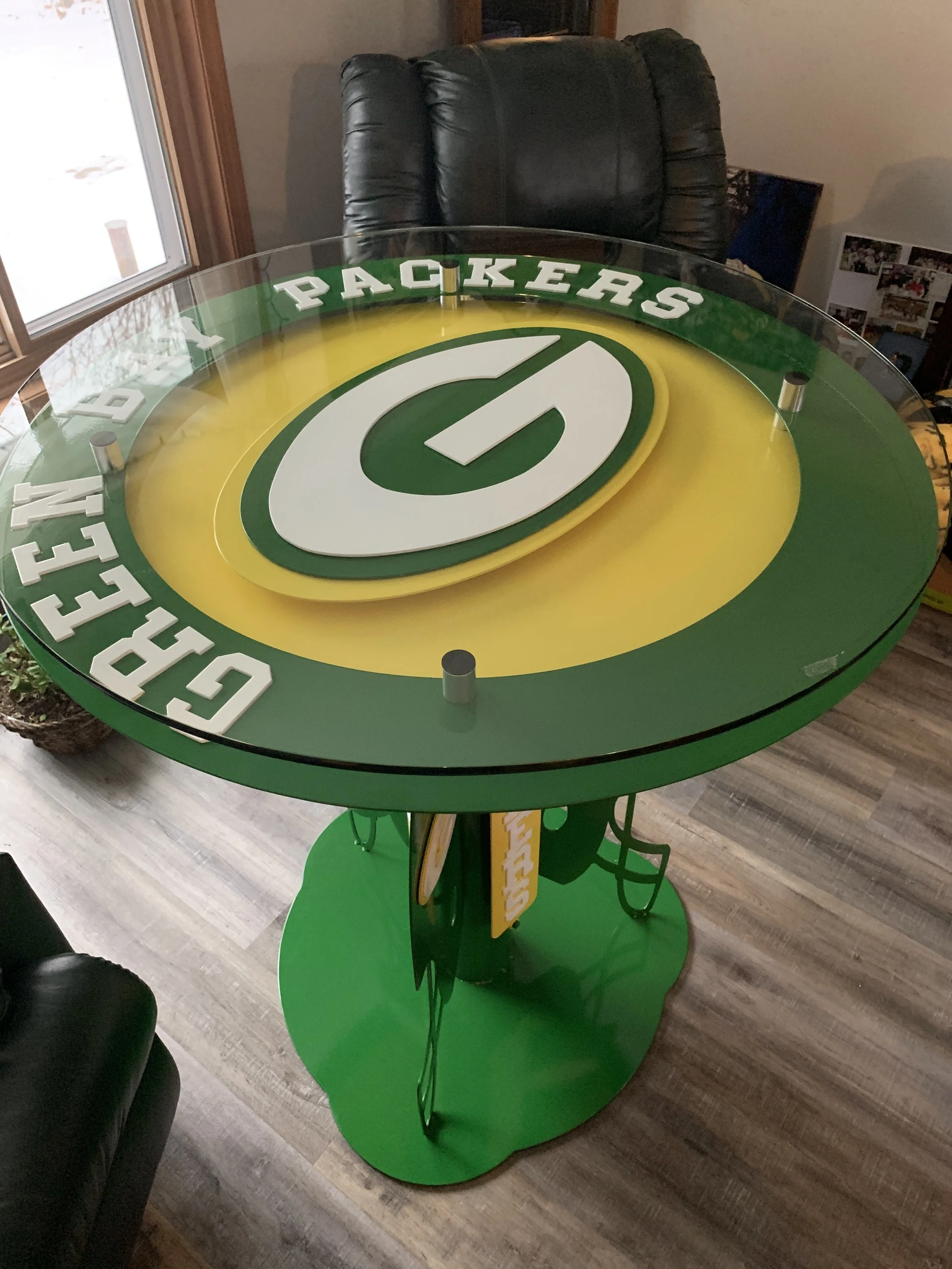Green and yellow Green Bay Packers-themed tabletop with a large Packers logo in the center, covered by a clear circular glass top, situated in a room with wooden flooring and a black leather chair behind it.