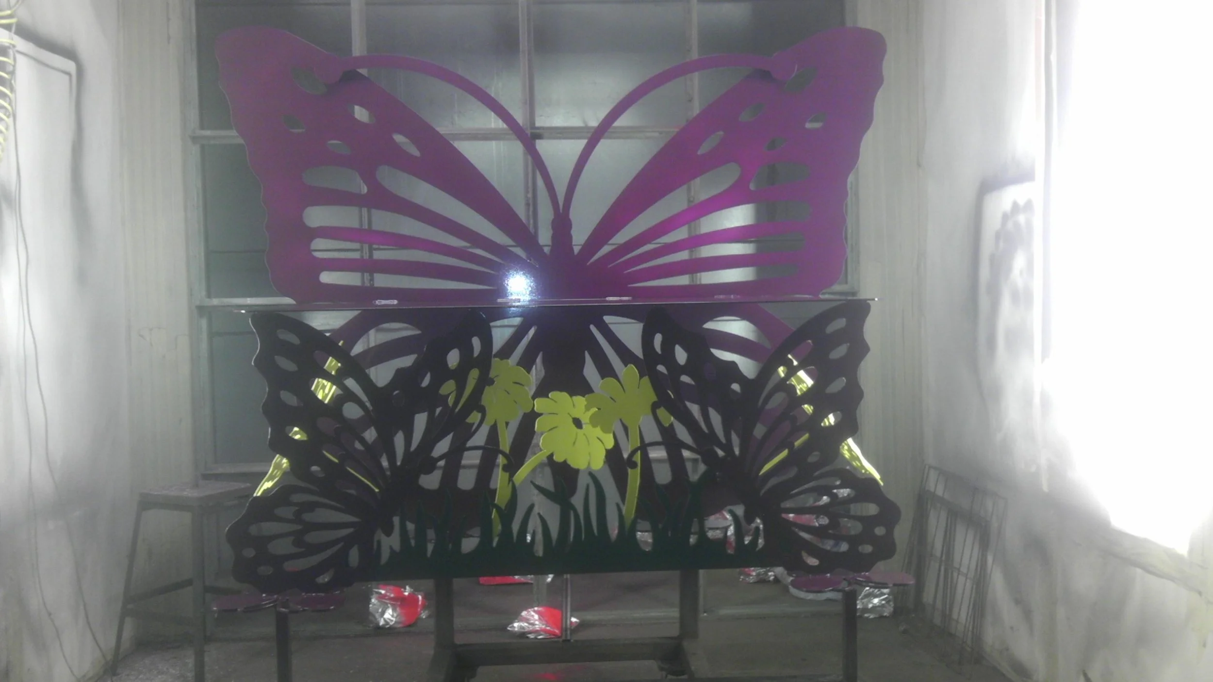 A decorative display of three large butterfly cutouts, purple on the top, black with yellow flowers on the bottom, mounted on a frame in a room with white walls and a window on the right.