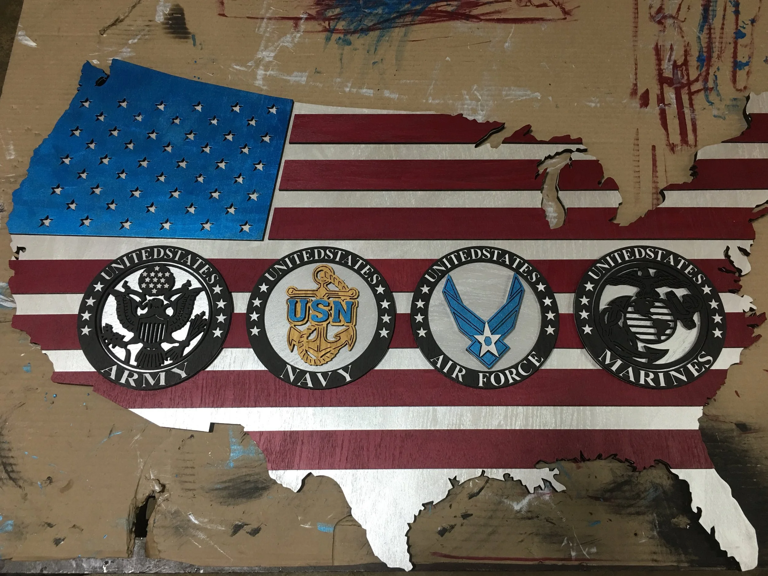 A wooden cutout of the United States map painted with the American flag, featuring the logos of the U.S. Army, Navy, Air Force, and Marines across the center.