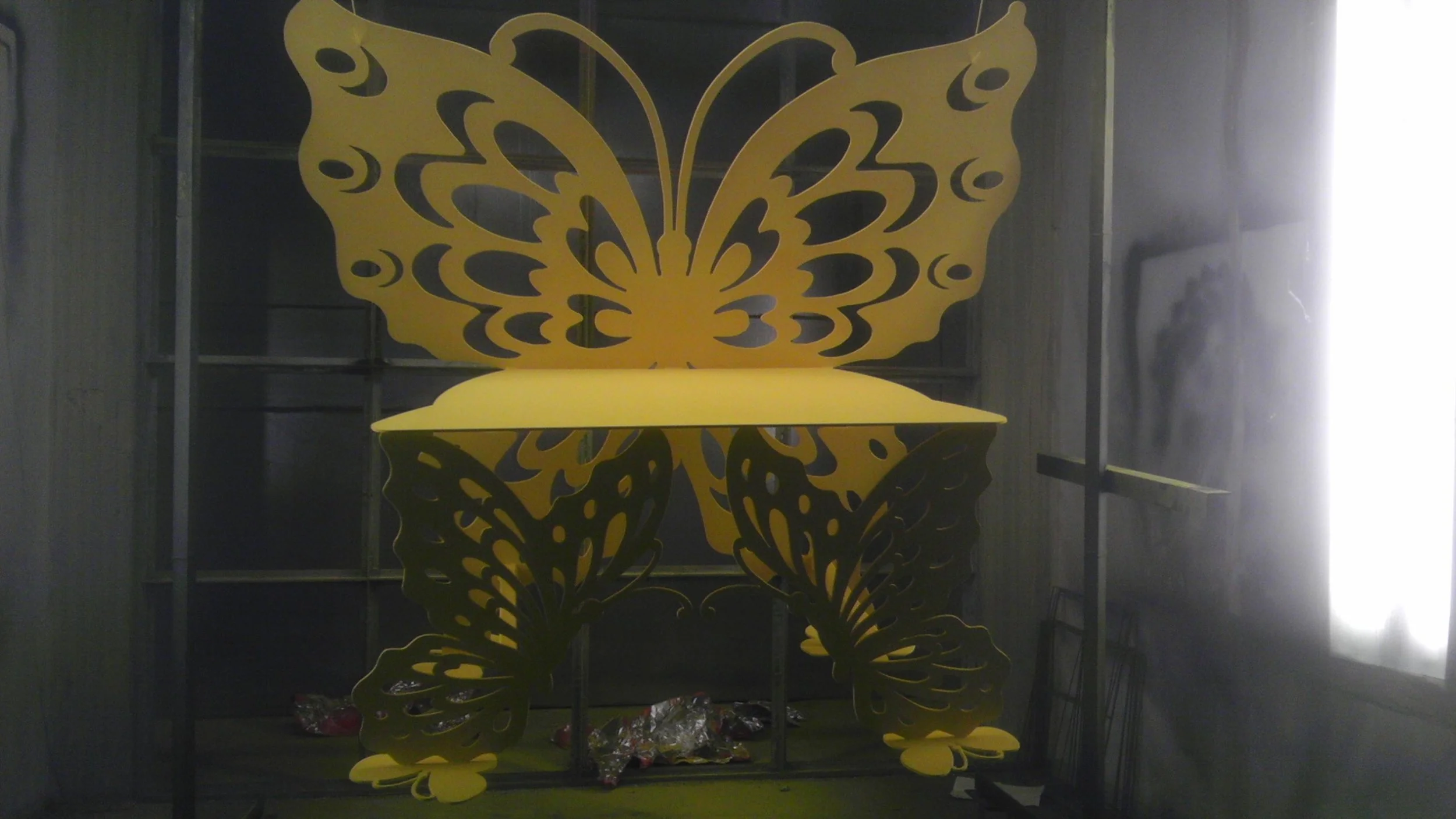 A large yellow butterfly-shaped structure with intricate cut-out details, sitting on a black metal frame, with a yellow platform on top, inside a dimly lit room.