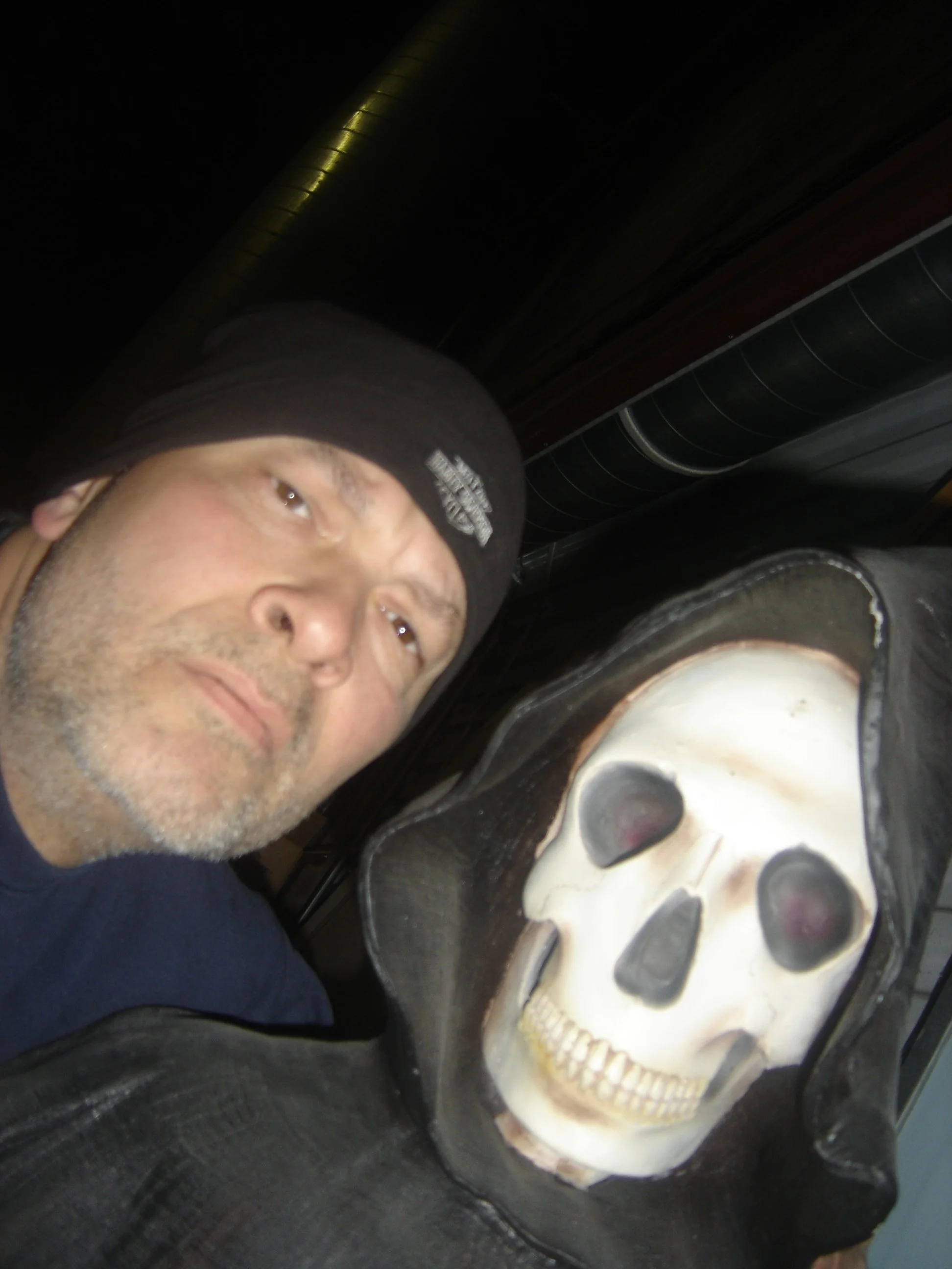 A man wearing a black beanie and dark clothing takes a selfie next to a large Halloween decoration of a skull with a hood.