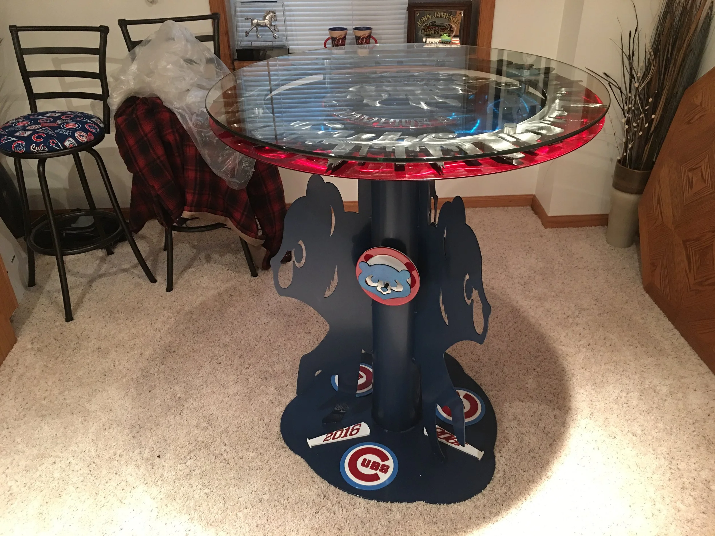A round glass-topped table with a Chicago Cubs themed base and decorations, situated in a room with beige carpet, surrounded by chairs and various decorative items.