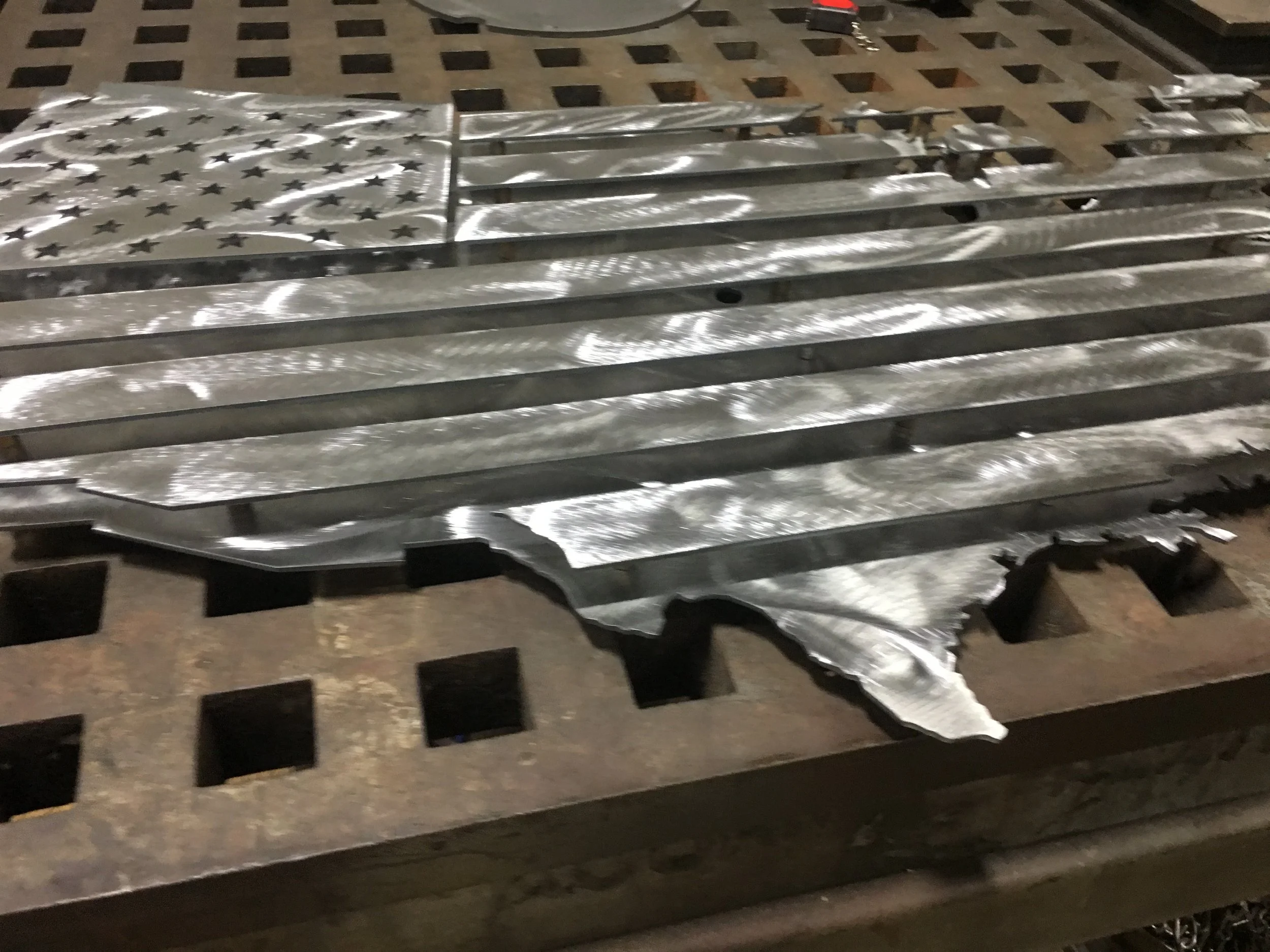 A metal sculpture of the United States flag with a damaged corner, lying on a metal grid work surface. The flag features stars on a blue field and horizontal stripes.