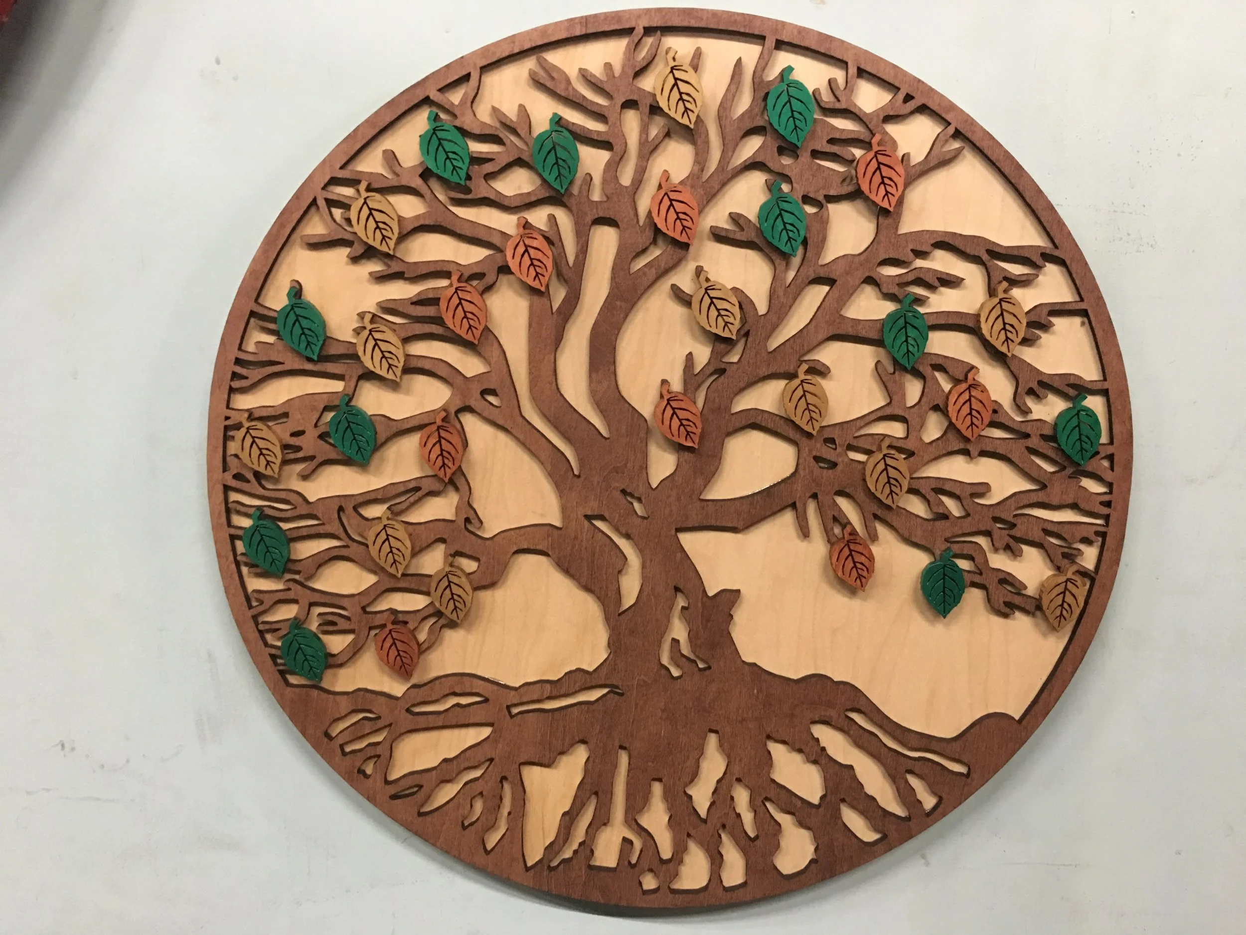 A round wooden wall art featuring a tree with branches, roots, and leaves in green, yellow, and orange colors.