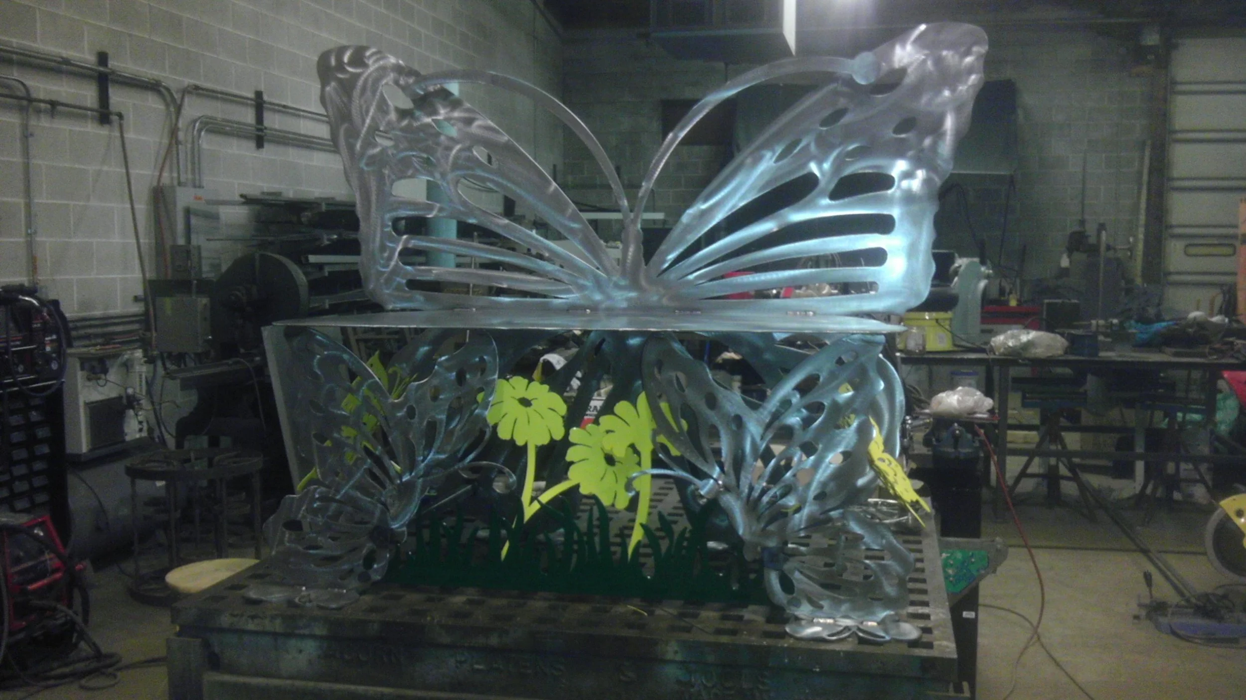 Large metal butterfly sculpture with illuminated wings, surrounded by yellow flowers, situated on a work table in a workshop.