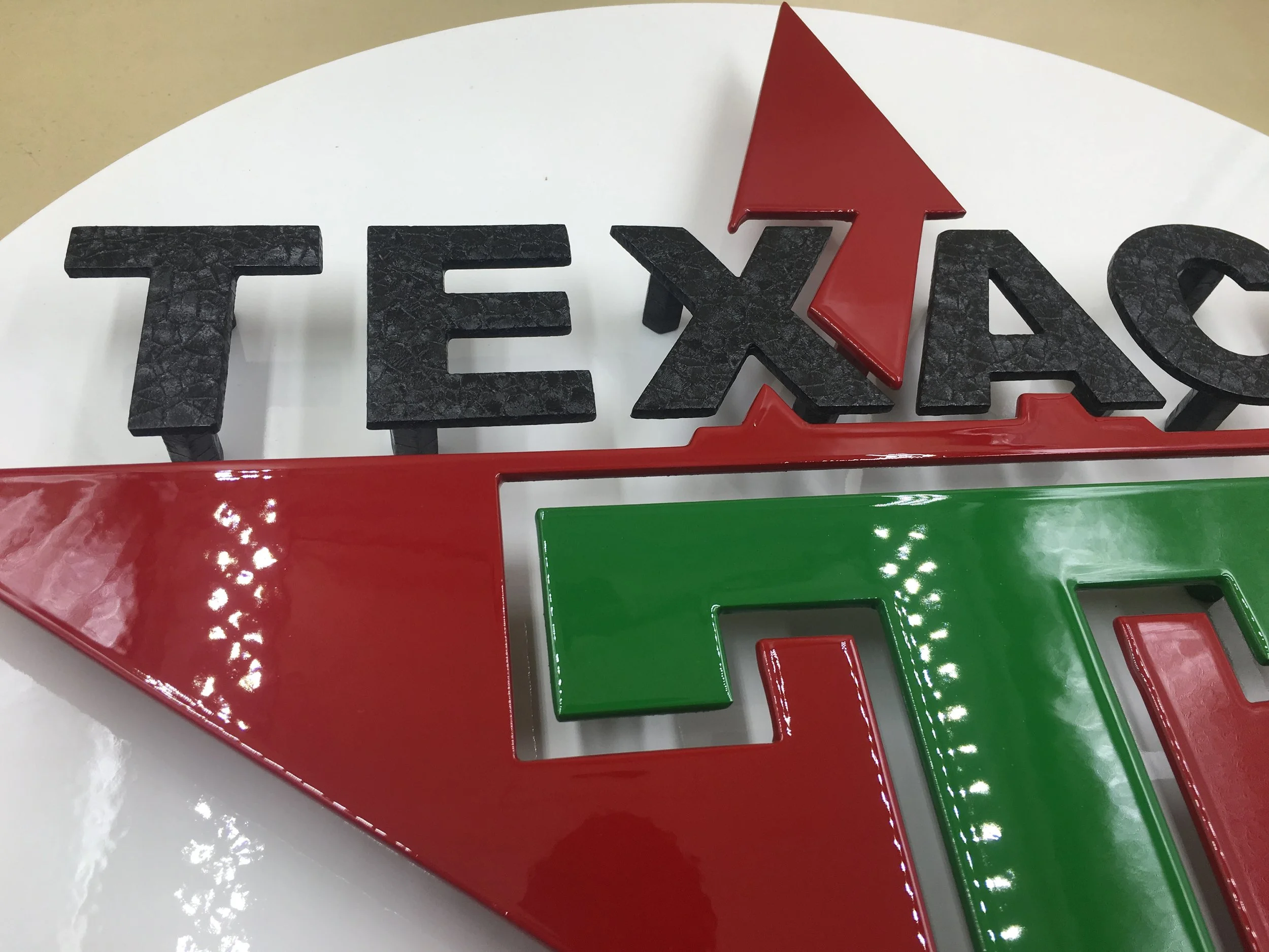 Close-up of a three-dimensional sign with the word "TEXAS" in black, textured letters and a red arrow pointing upward over the letter "X". The sign includes red and green decorative elements and is placed on a white surface.