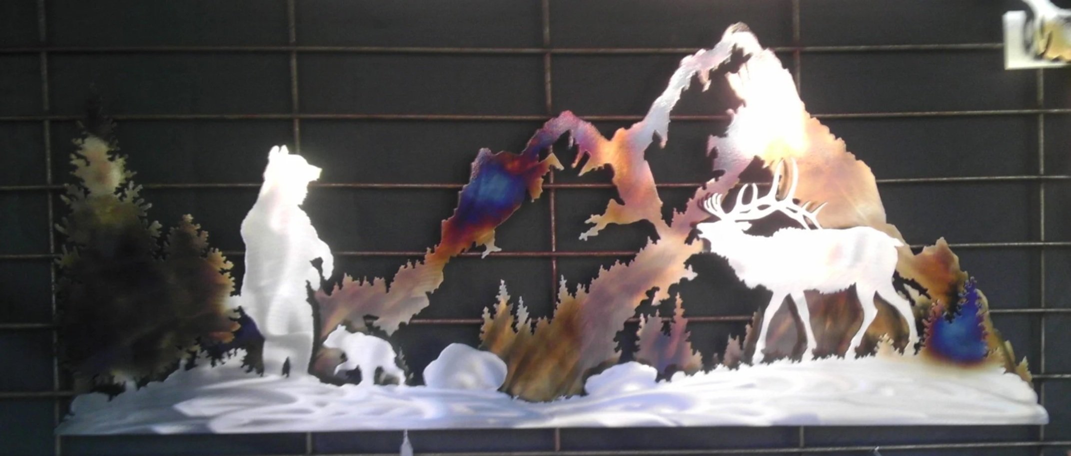 Metal artwork depicting a mountain scene with a reindeer, trees, and a person in a winter landscape.