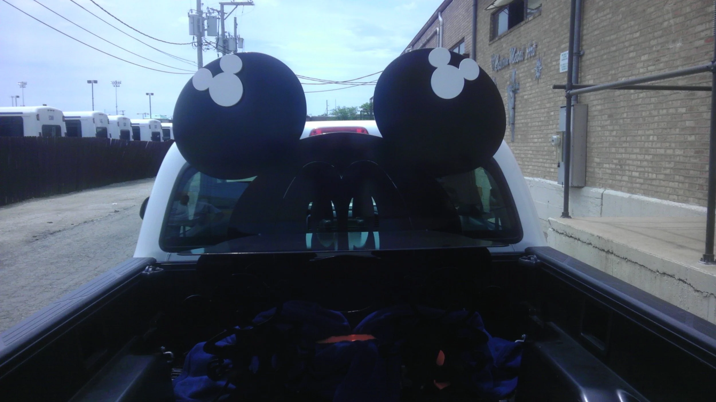 A vehicle with a large black Mickey Mouse ears headband on its roof, parked outdoors near a brick building and utility poles.