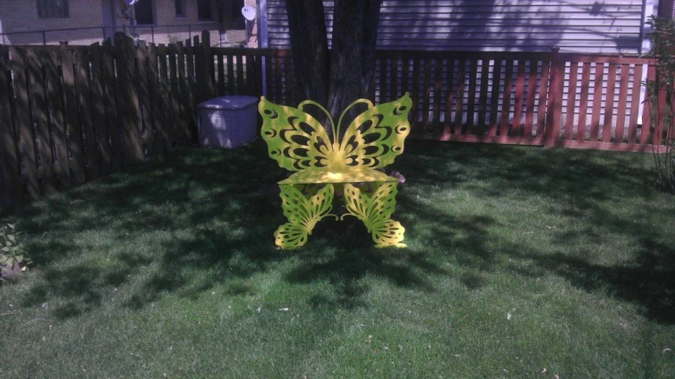Yellow butterfly-shaped garden bench in a backyard with green grass, fencing, and a trash can in the background.
