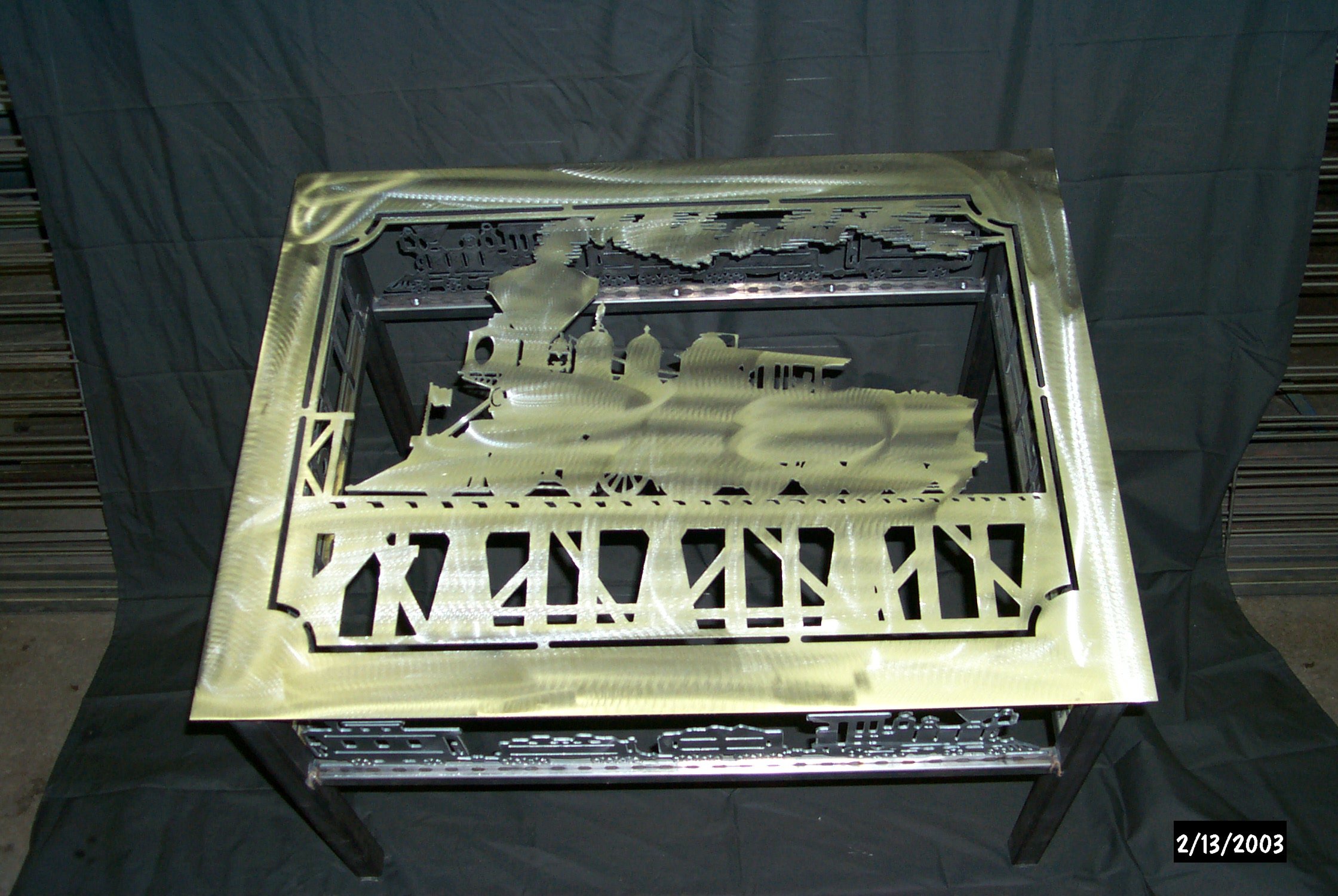 Metal cutout with the words 'Until We Meet Again' at the bottom, mounted on a display table. The cutout appears to be shaped like a train or machinery, with an industrial design.