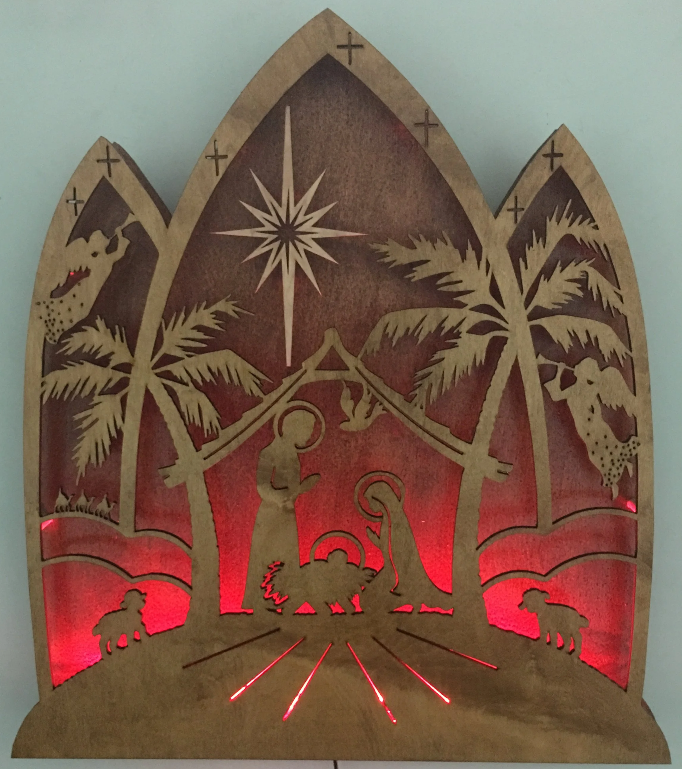 Wooden nativity scene with silhouettes of Mary, Joseph, baby Jesus, the star of Bethlehem, three wise men, sheep, and palm trees, illuminated with red lights.