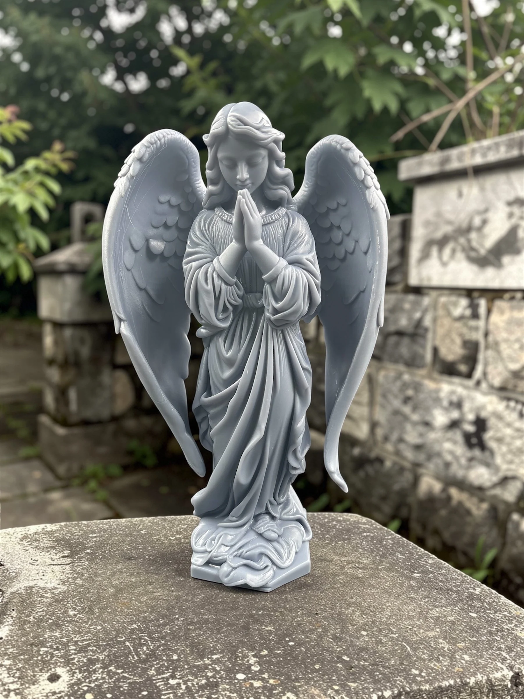 Gray angel statue with folded hands, standing outdoors on a concrete surface, with greenery and stone structures in the background.