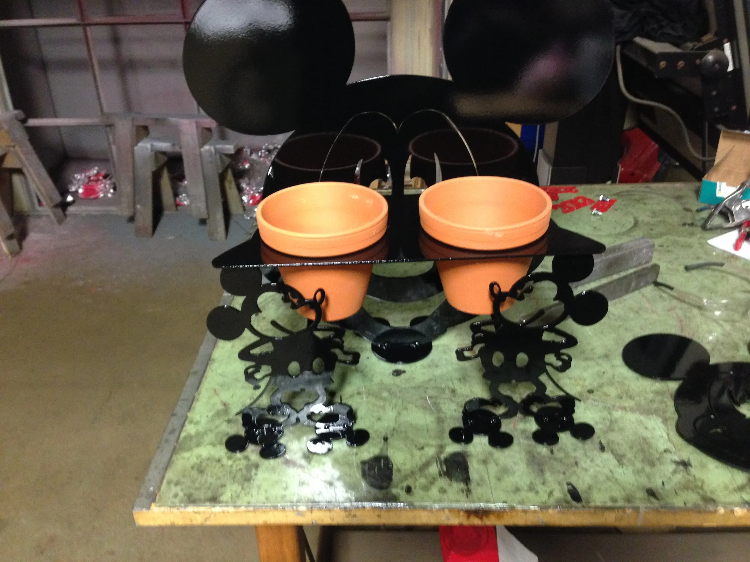 A black and orange Mickey Mouse-themed metal decorative piece with two small orange pots, placed on a green workbench with tools and metal parts nearby, in a workshop or storage area.