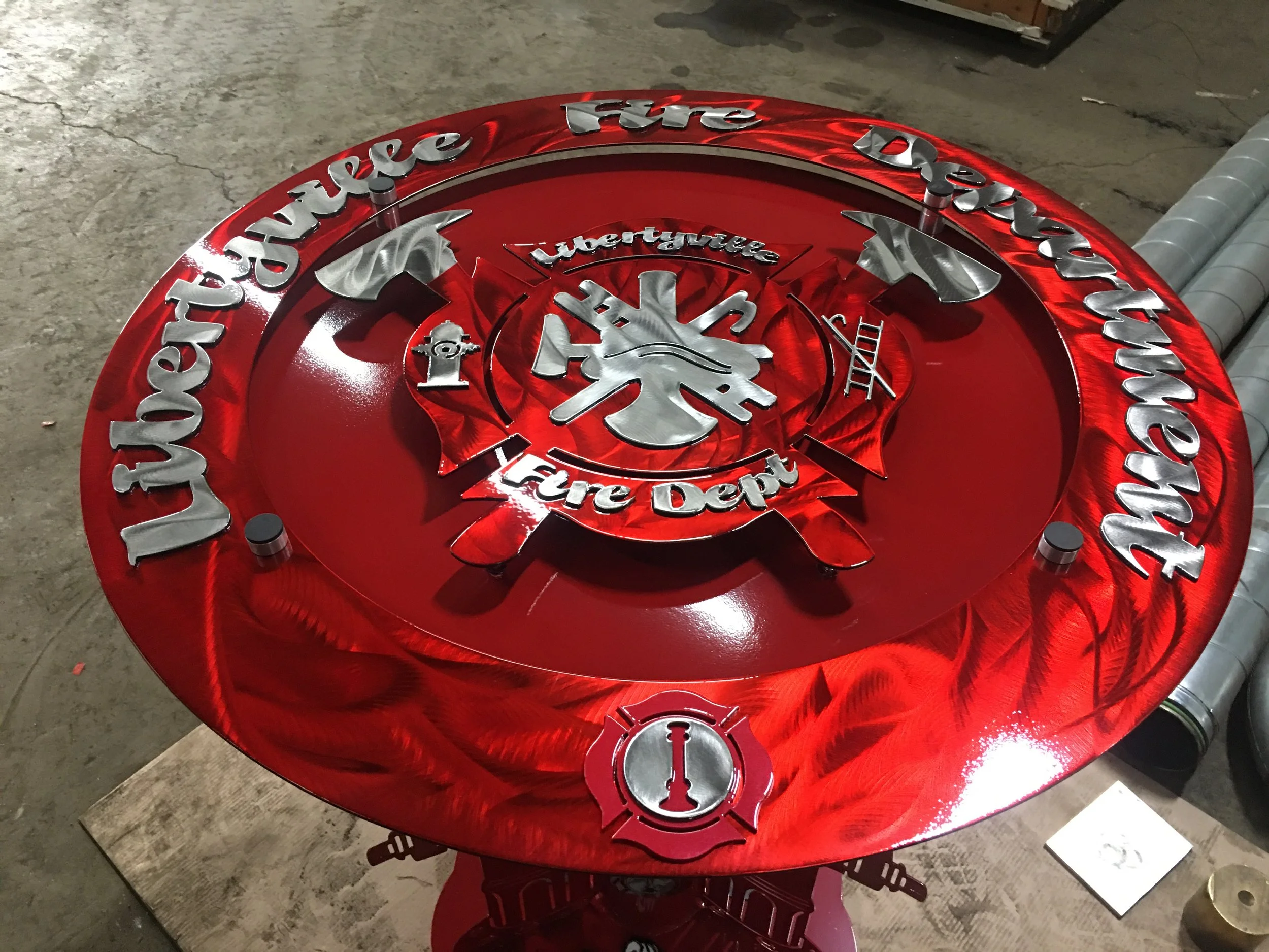 A large circular metal sign painted red with silver lettering and designs. It reads 'Wichita Fire Department' around the outer edge, with a fire department emblem in the center, including a fire hydrant and crossed axes.