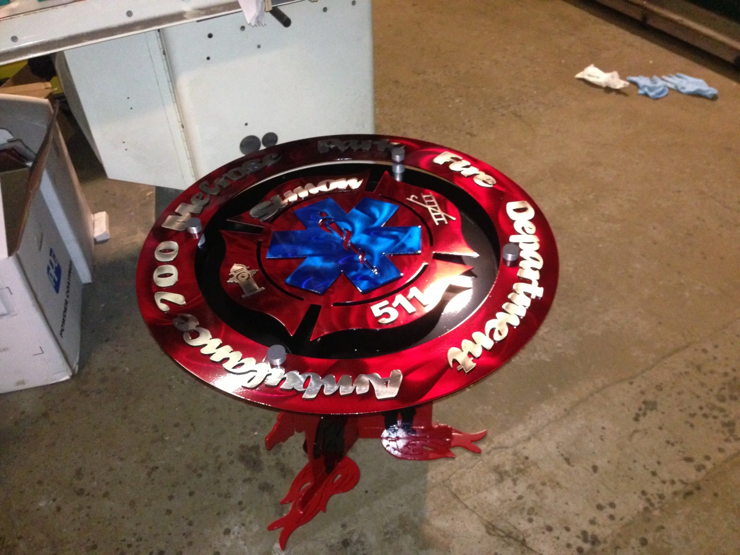 A circular outdoor decoration featuring the number 911, with a blue star of life symbol at the center, and emergency services references including the American flag, the words "Ambulance," "Fire," and "Rescue" in metallic lettering, and contact number 700, on a concrete ground.