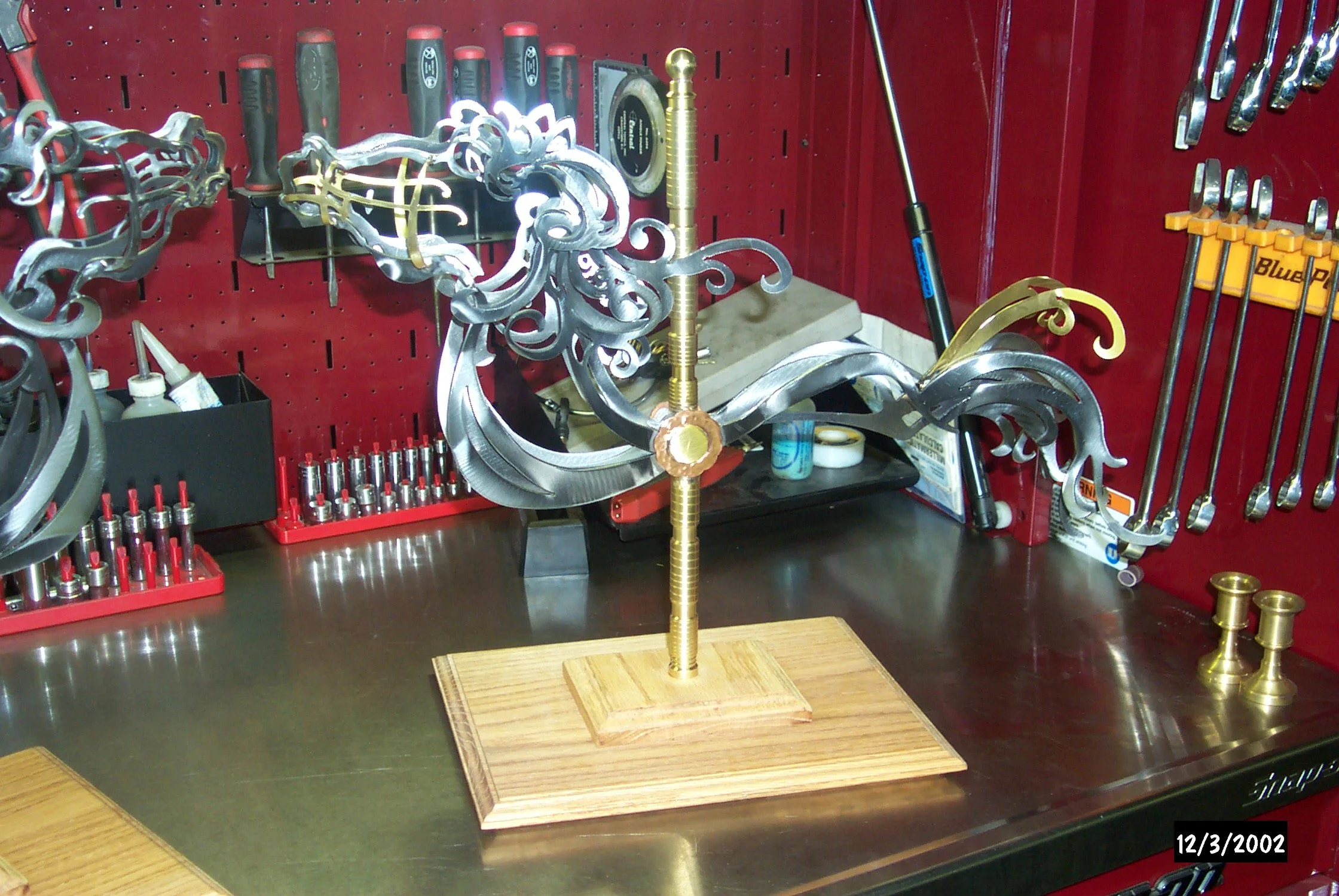 A metal sculpture resembling a dragon or creature with ornate, curving silver and gold details mounted on a wooden base on a workbench in a workshop.