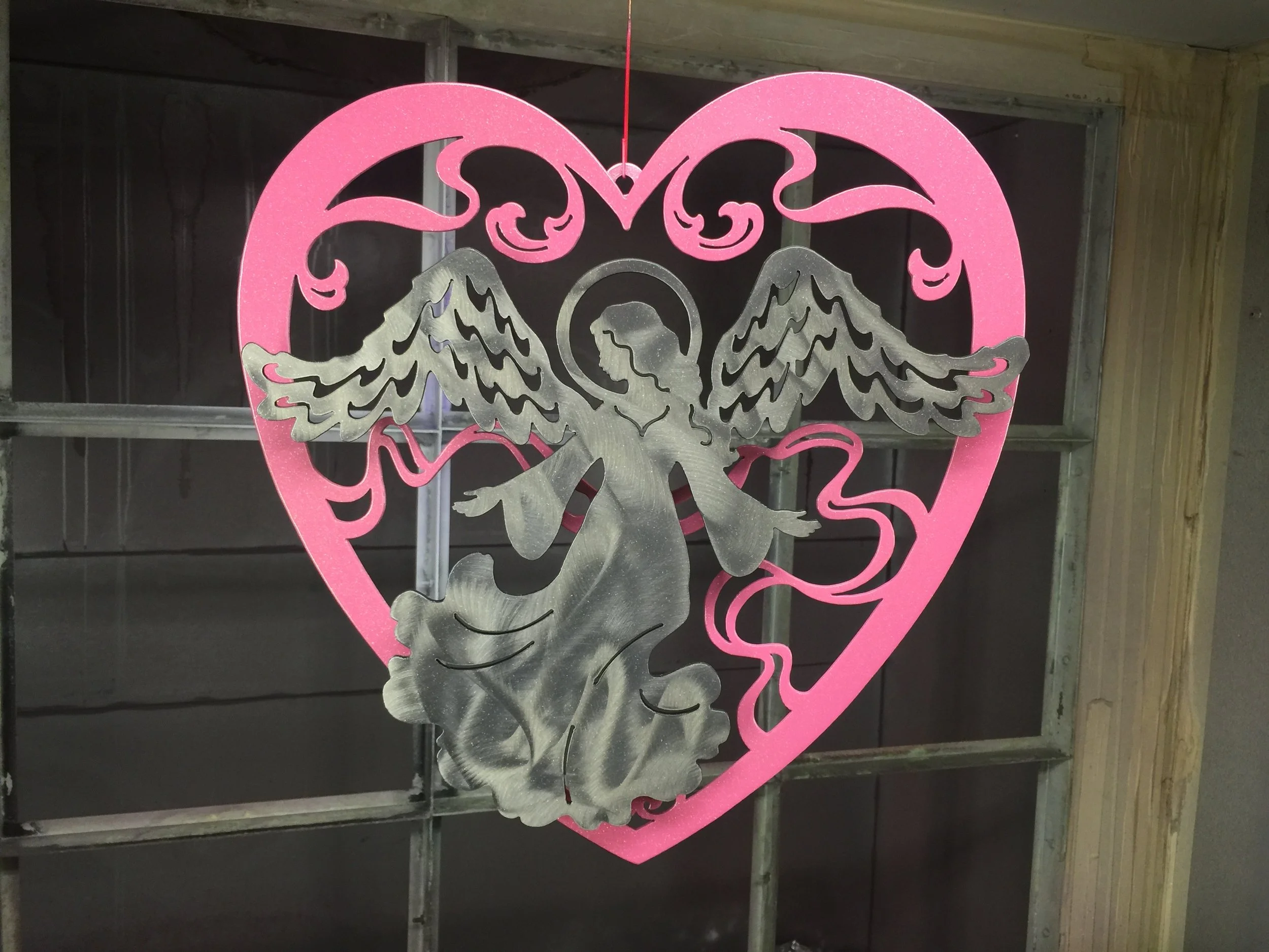 A decorative hanging ornament featuring a silver angel with wings inside a pink heart outline, against a background of construction or unfinished room.