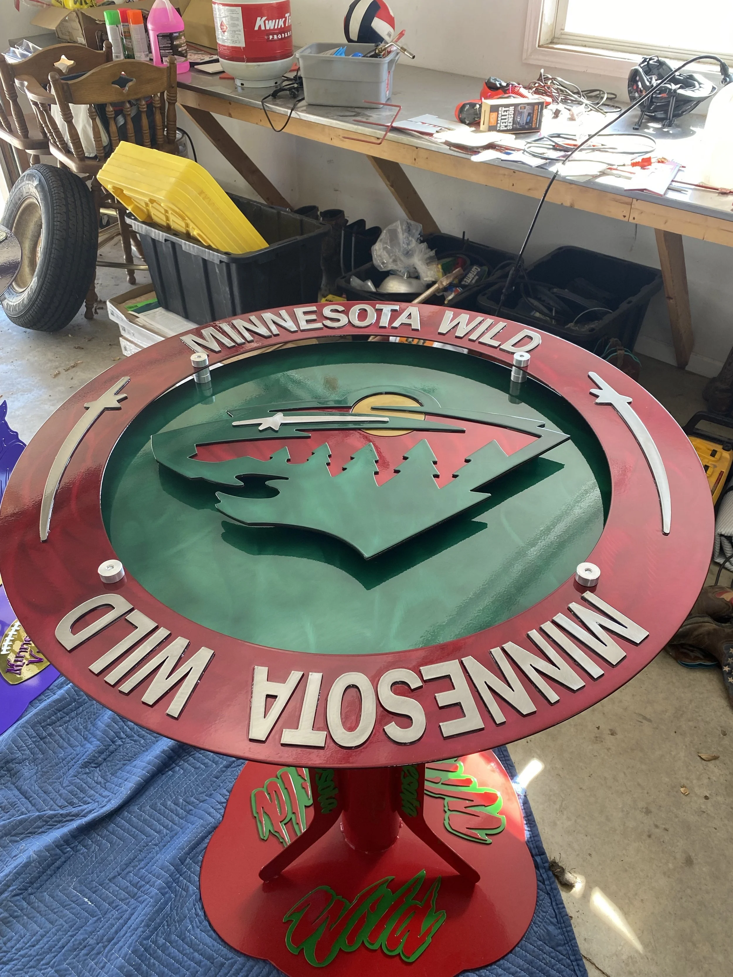 A circular Minnesota Vikings-themed clock with a tree and mountain design, red outer rim, and green face, placed on a table in a workshop.