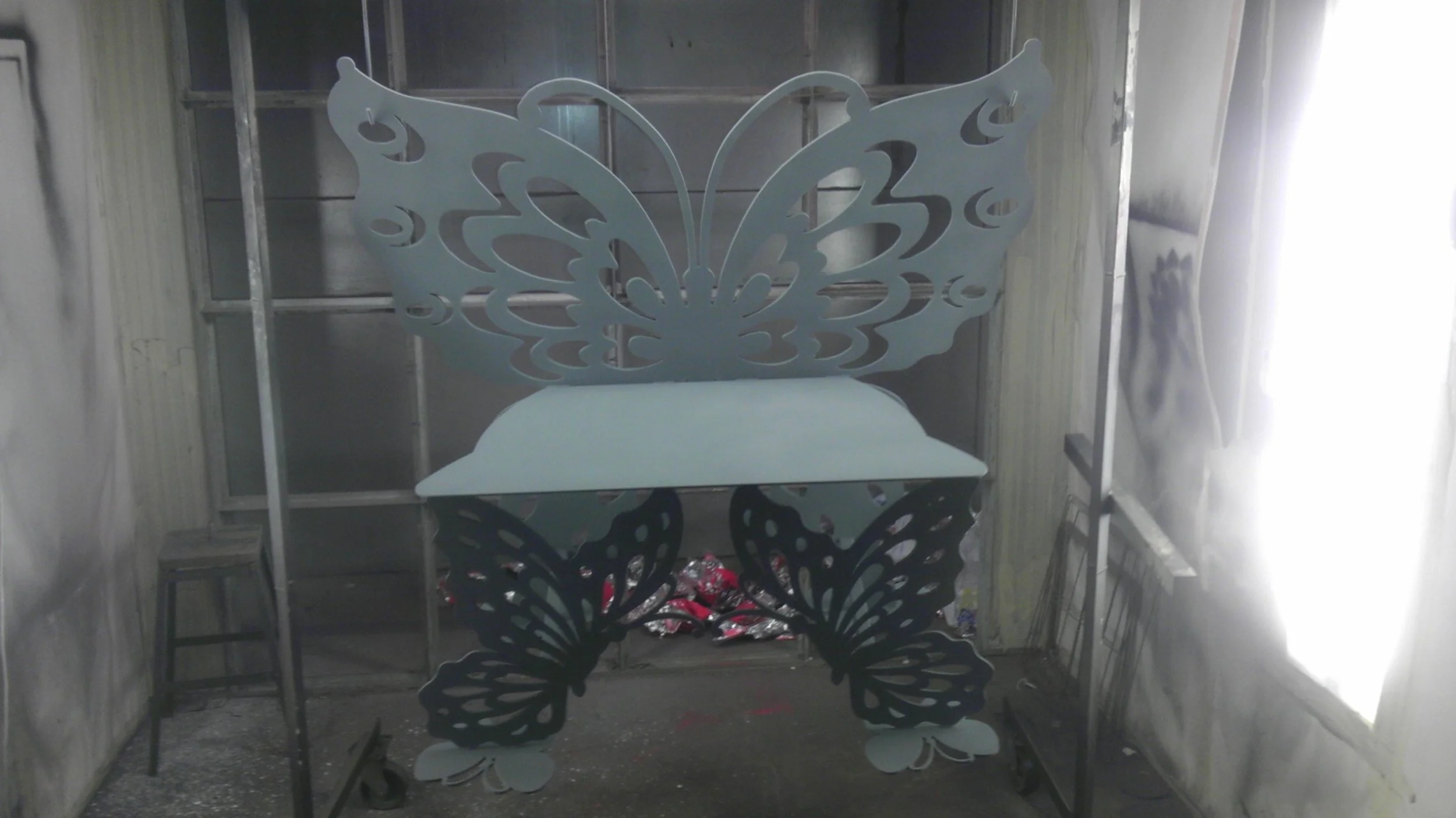 A large butterfly-shaped metal display with wings and legs, painted in white, standing on two black butterfly-shaped legs inside a workshop or industrial space.