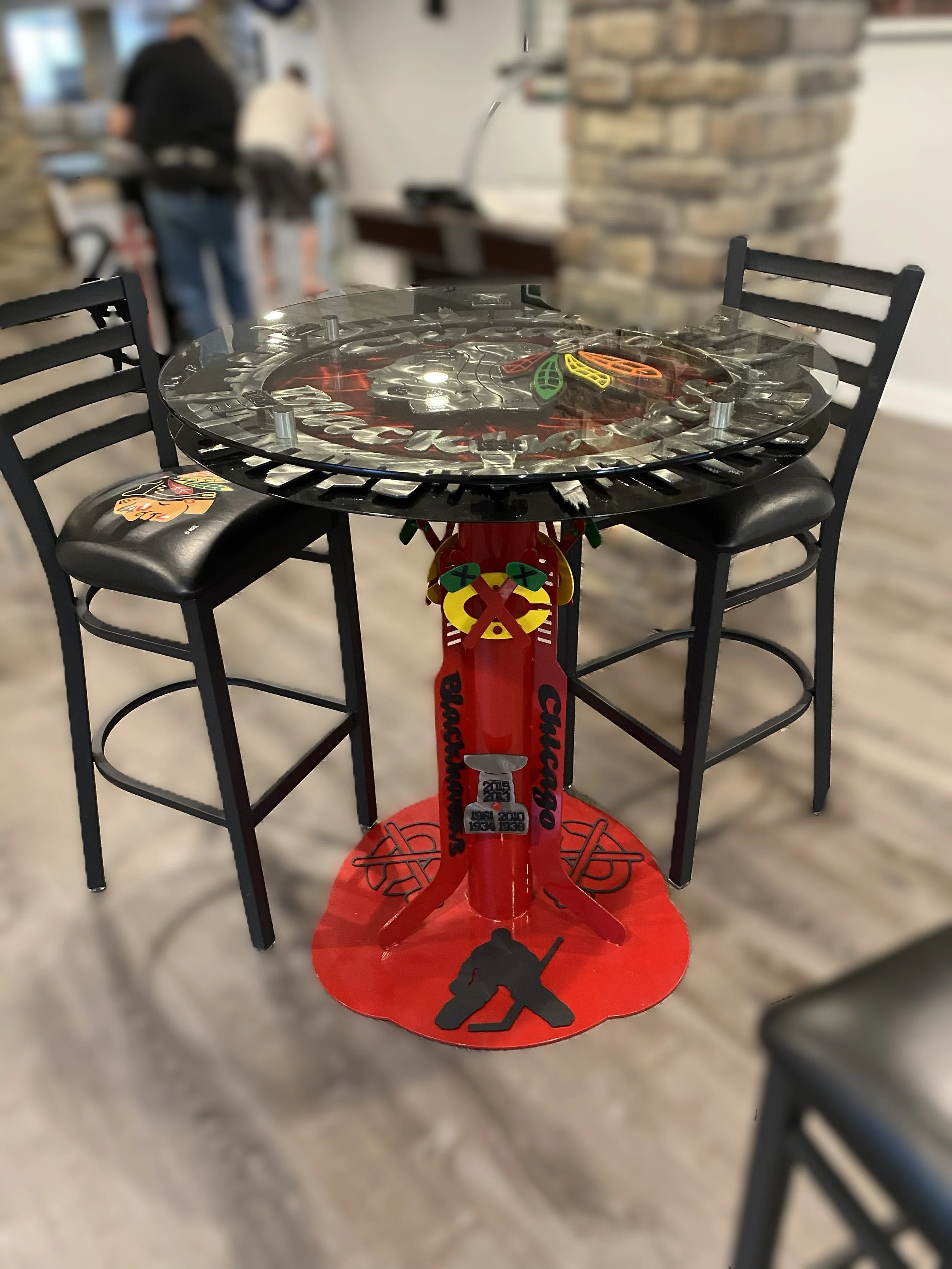 A round metal table with a glass top featuring the Chicago Blackhawks logo, surrounded by black chairs, with indoor restaurant or cafe background including a brick column and blurred people.