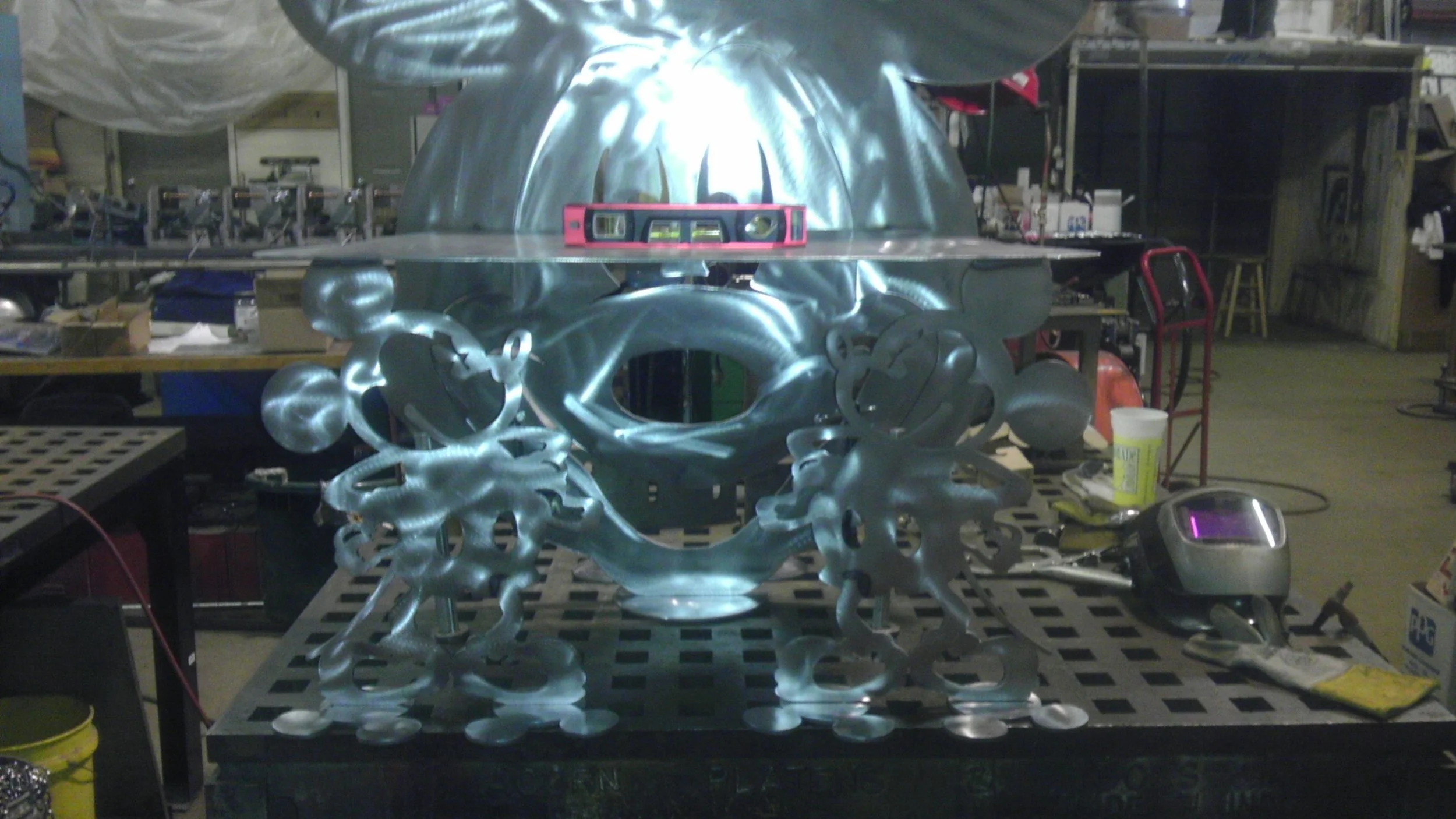 Metal sculpture resembling a face with large earrings, situated on a work table in a workshop environment, with various tools and equipment around.