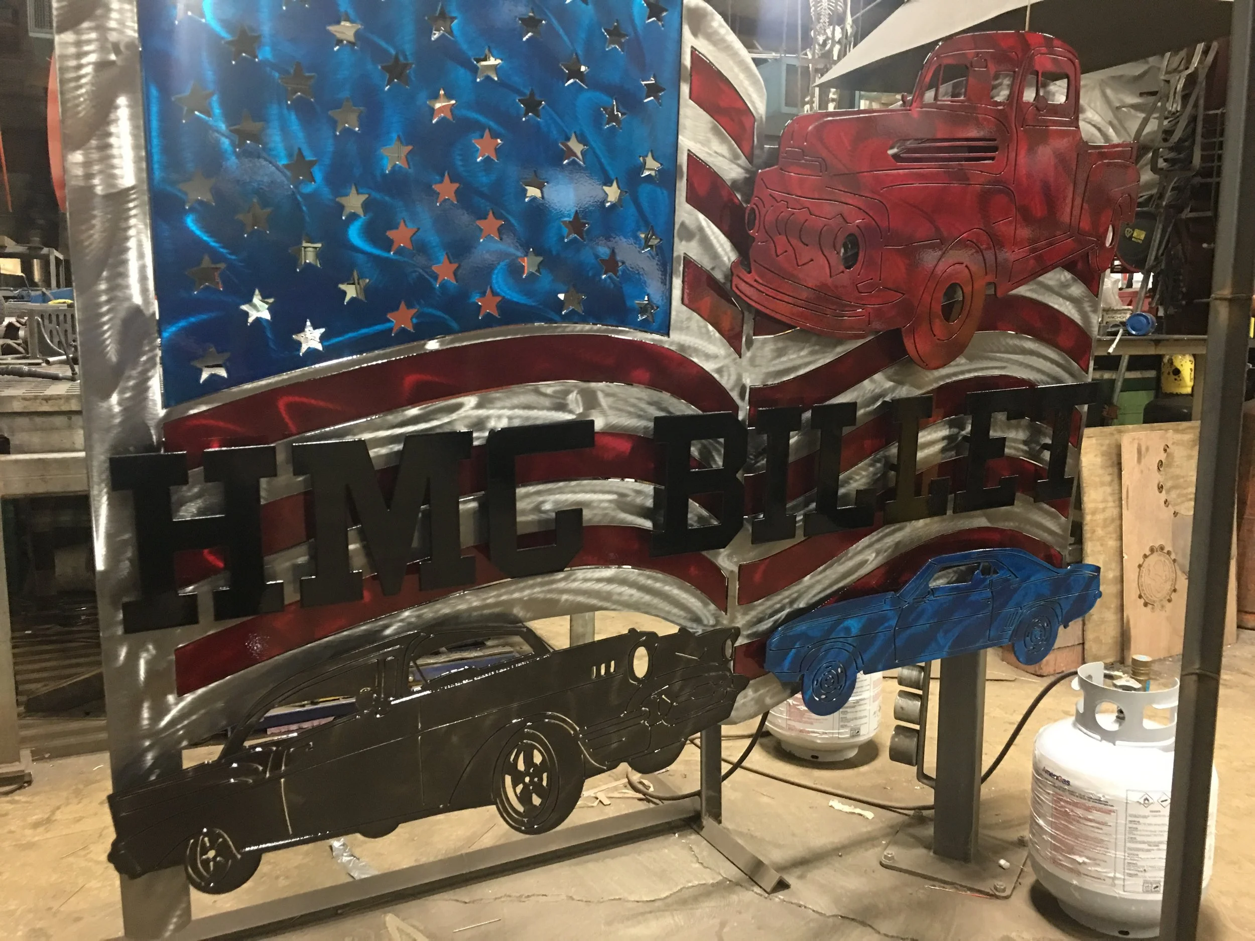 Decorative metal sign with patriotic theme featuring stars, stripes, and images of vintage trucks, with the word 'HMC BILLE' in bold black letters.