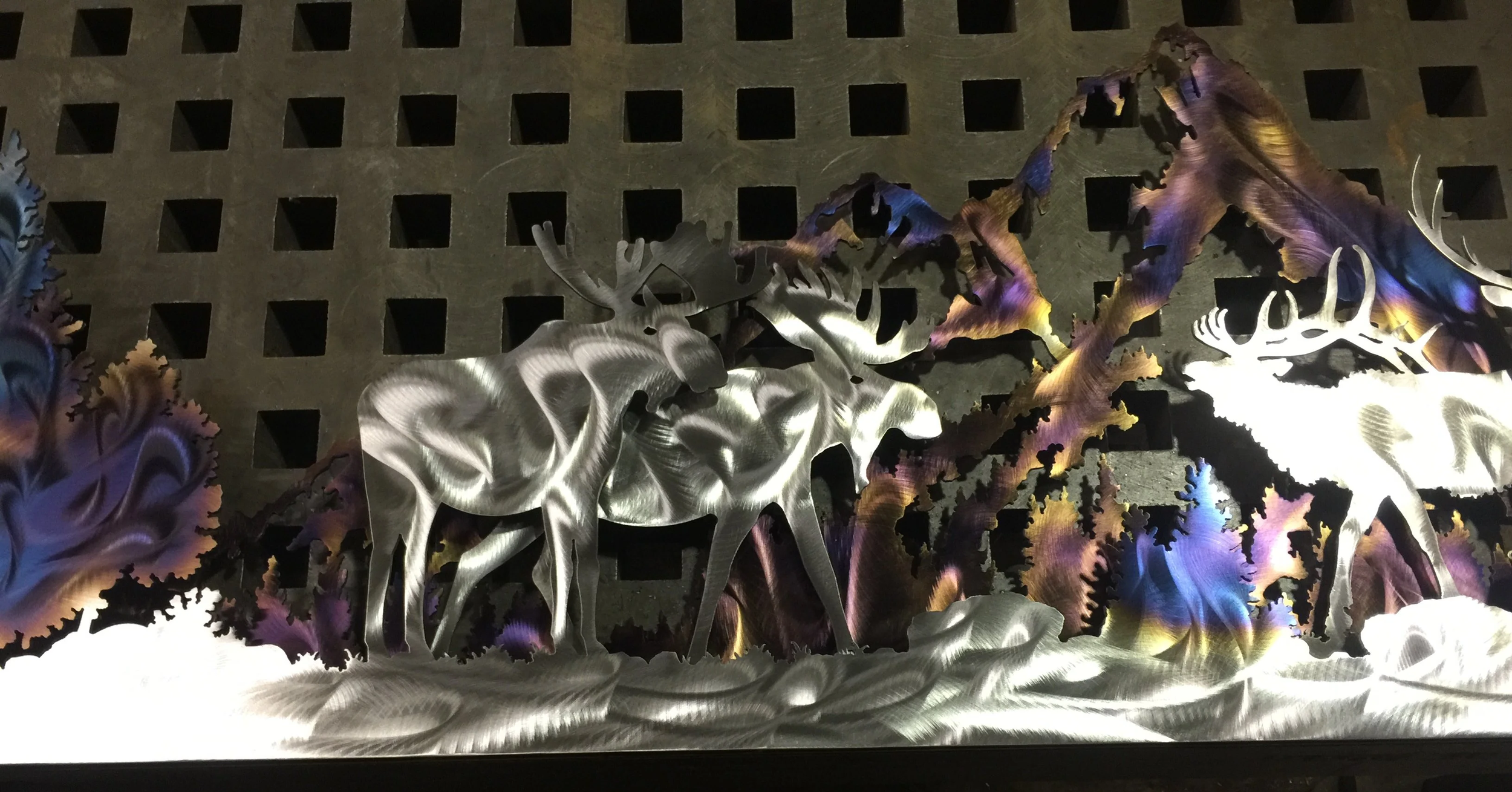 Metal wall art depicting a moose standing in front of trees, with colorful reflections on the metal surface.