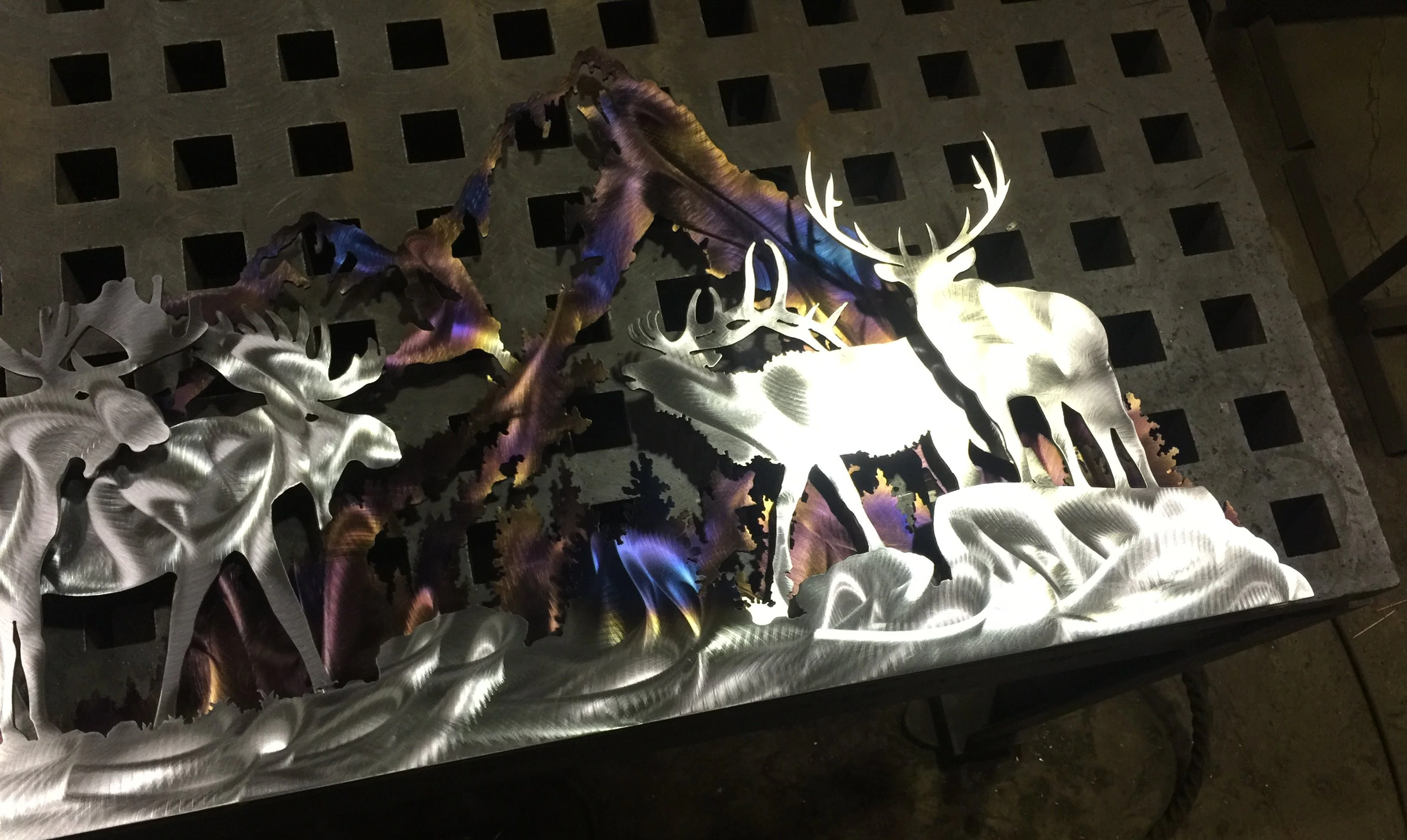 Metal wall art depicting a silhouette of a deer with antlers, surrounded by trees and foliage, illuminated by colorful lighting.