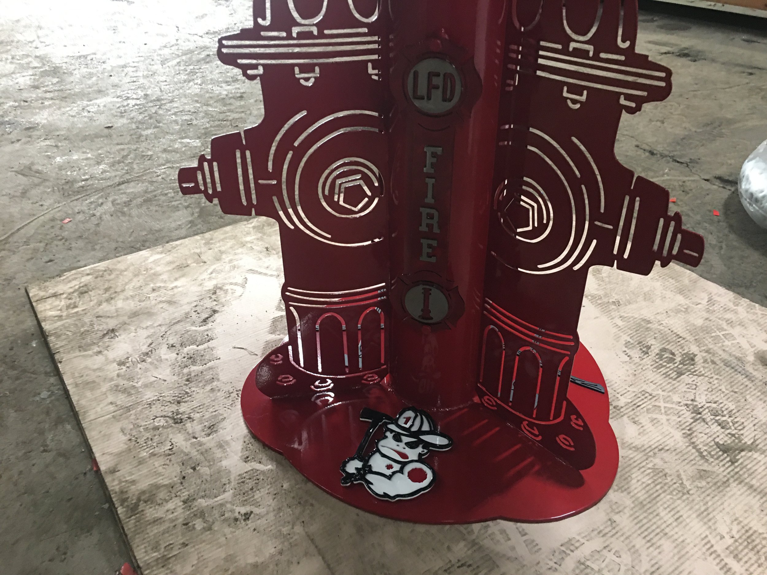 A red metal fire hydrant-shaped stand with a cartoon mouse holding a golf club logo at the base and the words 'fire' and 'fire dept' on it. The stand is on a wooden plank on a concrete floor.