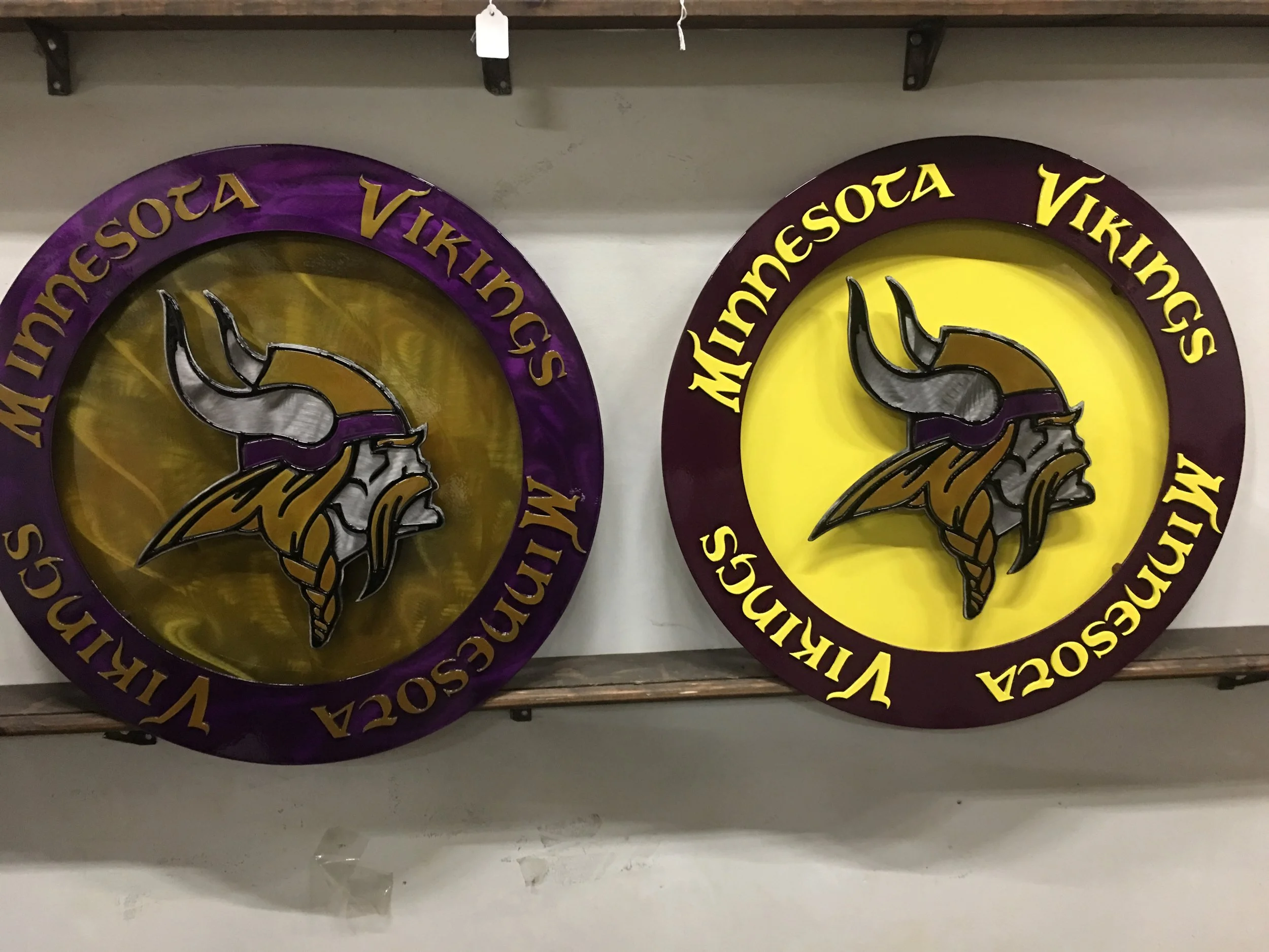 Two Minnesota Vikings logo signs side by side, one with a purple border and yellow background, the other with a maroon border and yellow background, featuring a Viking head with a helmet and horns.