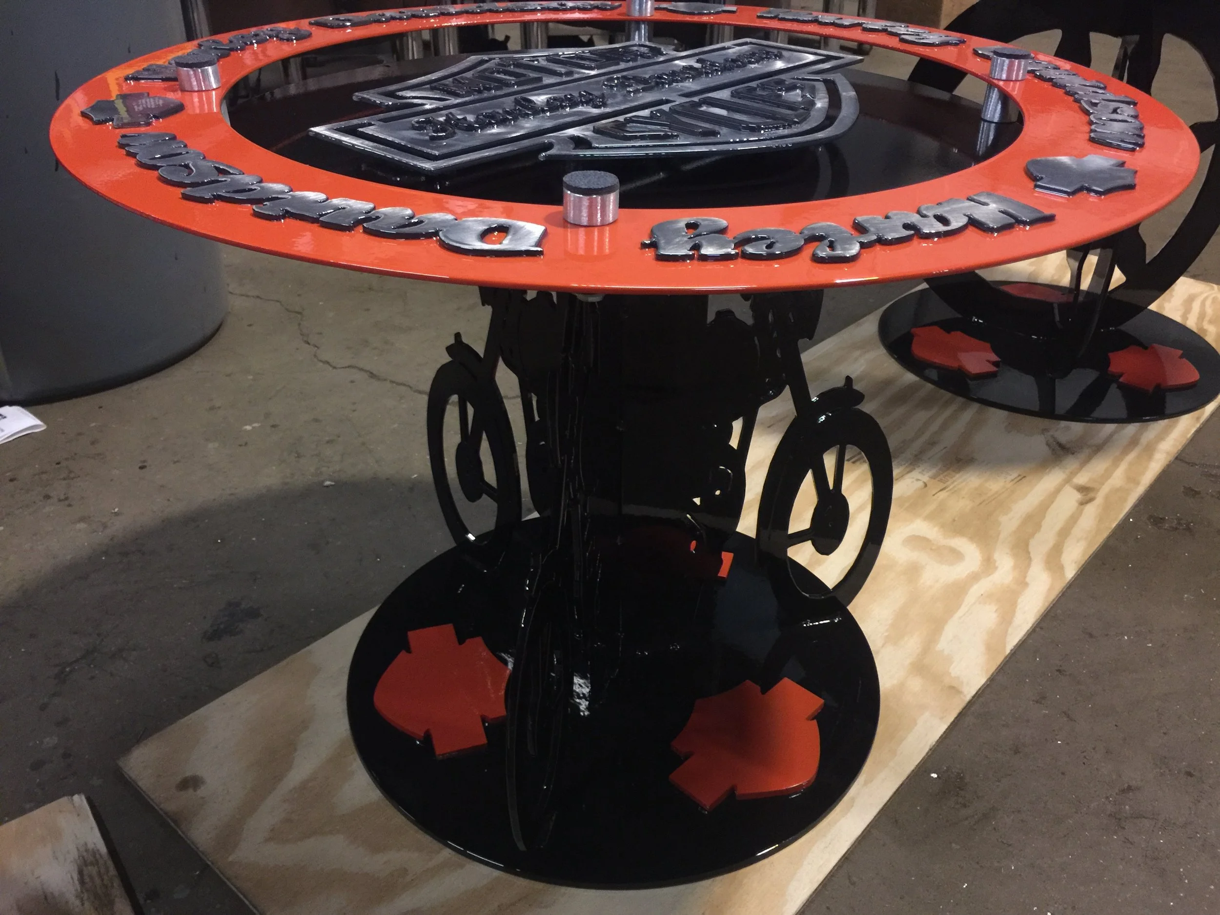 A round table with a black top and a decorative leg base, featuring a red border with black raised letters spelling 'Marijuana Reception' and medical symbols, located on a wooden platform in a warehouse or workshop setting.