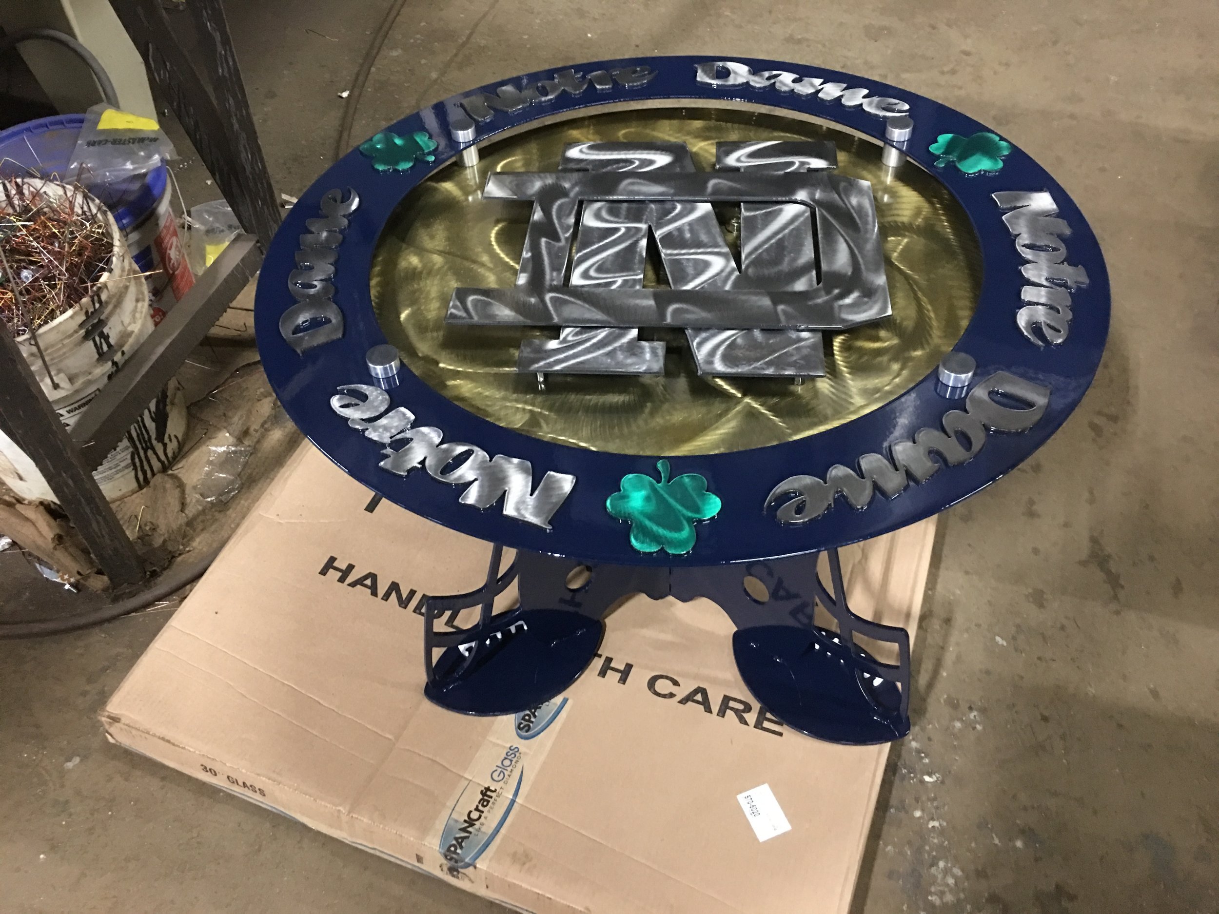 St. Patrick's Day themed metal table with a round blue border, shamrock decorations, and the words "Merry Christmas" and "Happy New Year" inscribed on it. The table has an iron central design and stands on a cardboard base in a workshop setting.