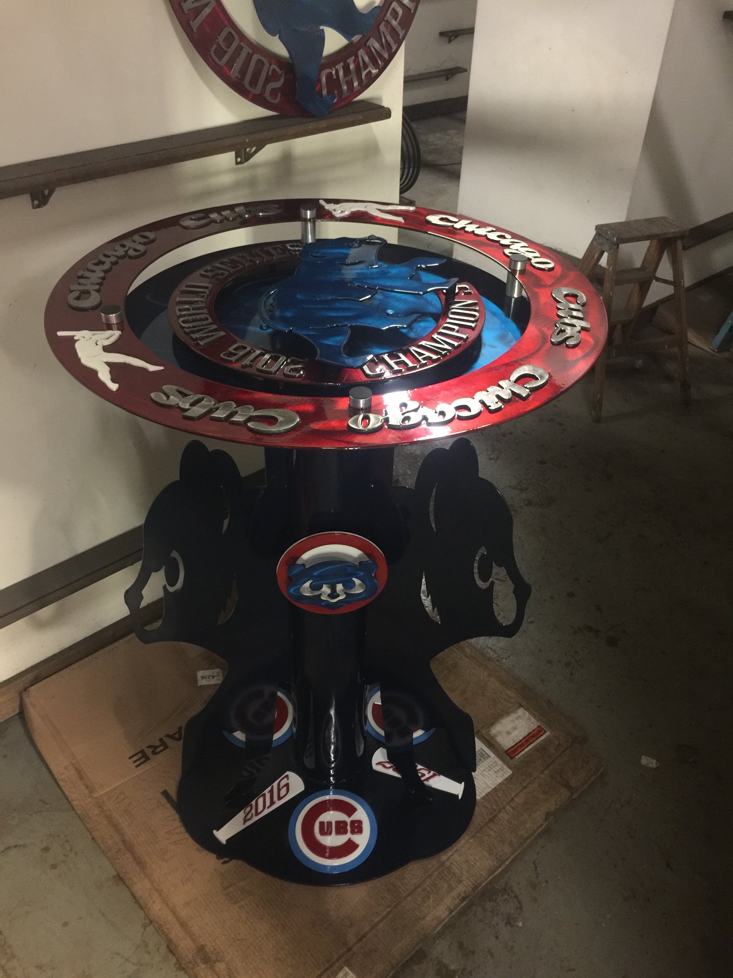 A Chicago Cubs-themed tabletop shuffleboard game with red, black, and blue colors and the Cubs logo, placed on a cardboard box in a room with wooden stools and a metal shelf.