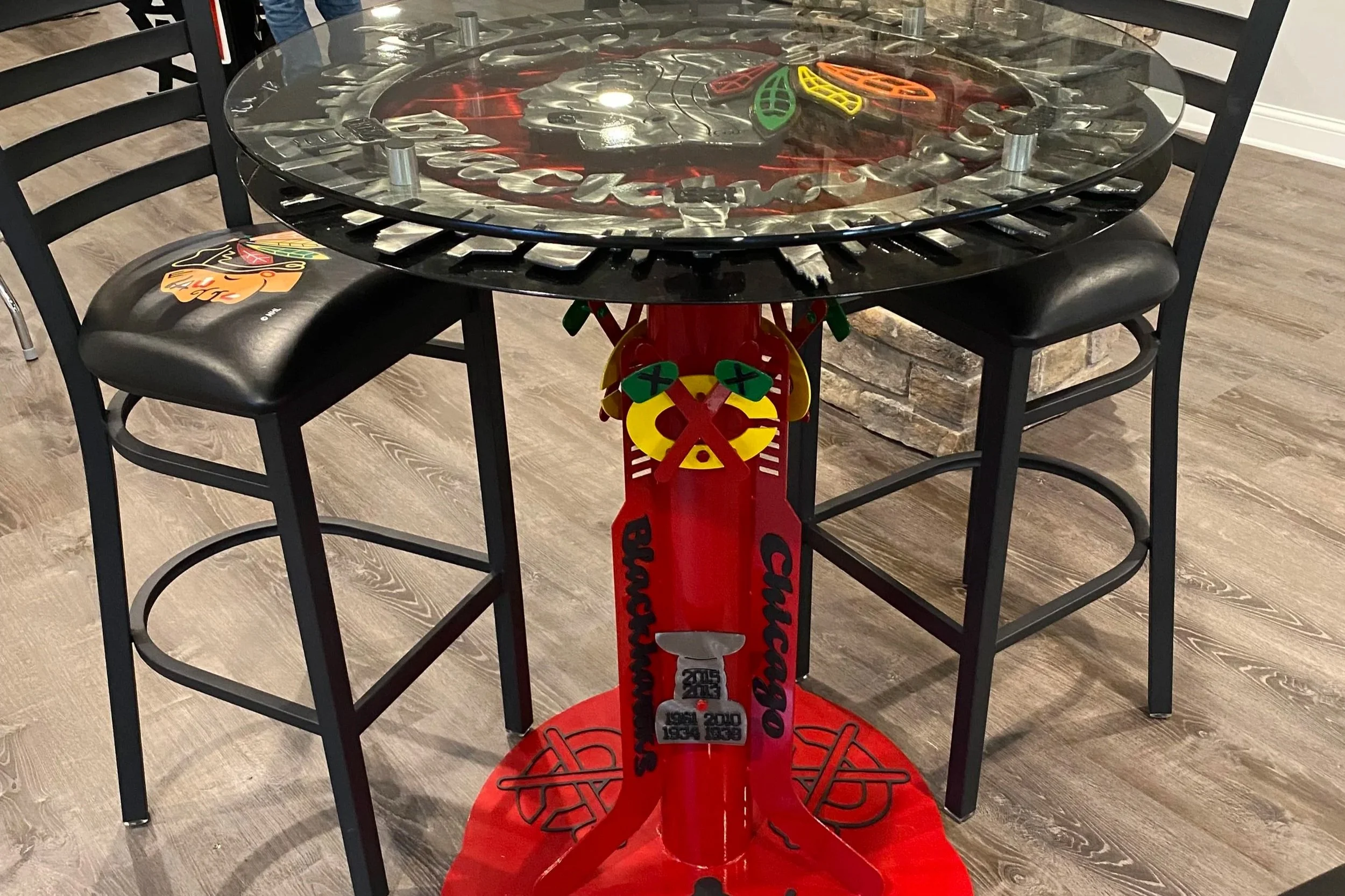 A round table with a glass top featuring a Chicago Blackhawks logo, surrounded by three black chairs, and decorated with a Chicago Blackhawks theme including a red stand with team graphics and a Stanley Cup image.