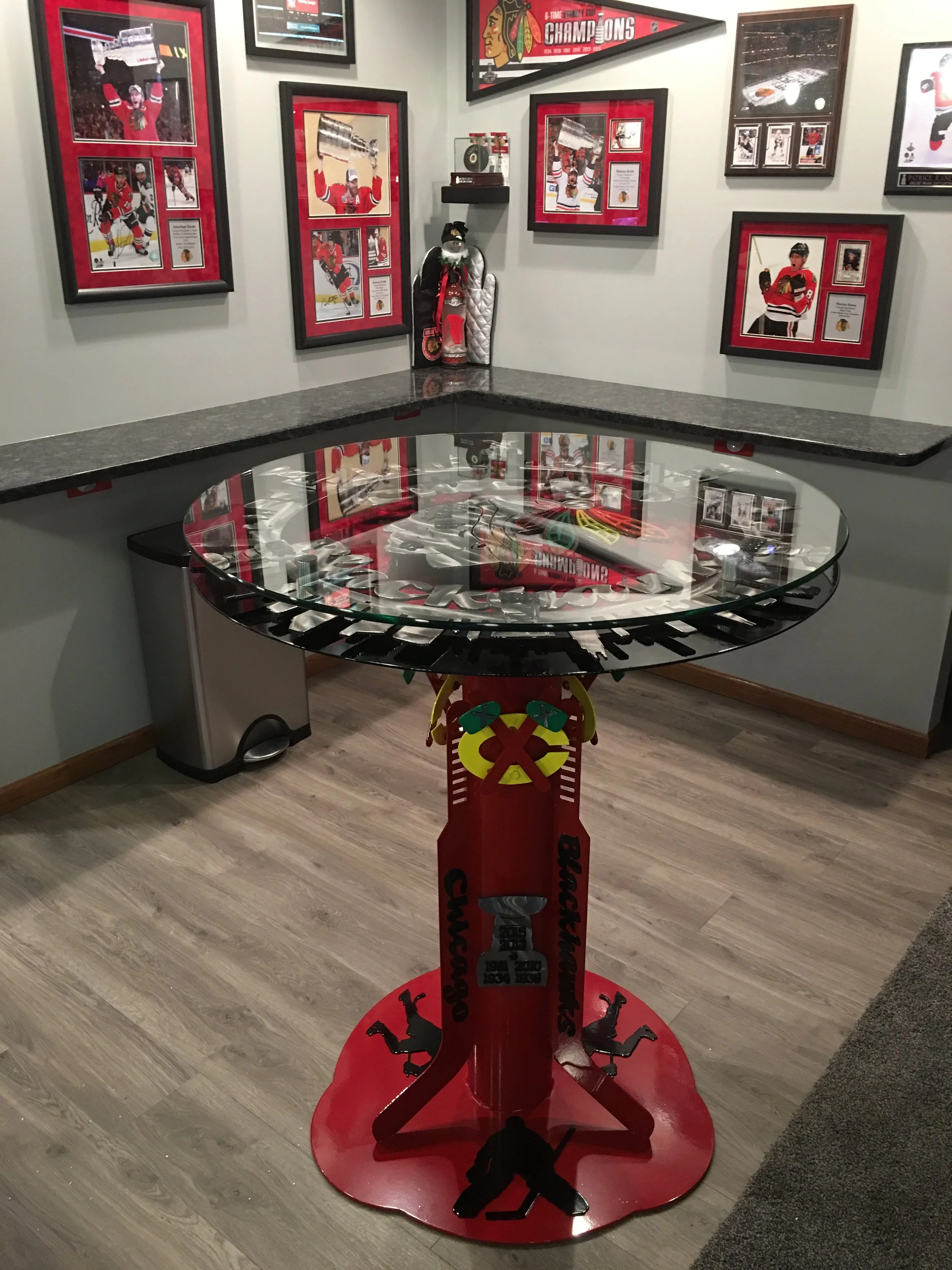 A hockey-themed display room with framed pictures and memorabilia. There is a round glass table with a hockey stick motif, a table base decorated with hockey players, and walls adorned with photos of hockey players, trophies, and team memorabilia, all celebrating the Chicago Blackhawks.