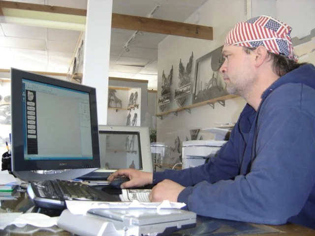 A man wearing a bandana with American flag design working on a computer in an office or workshop with mounted decorative metal art and shelves in the background.