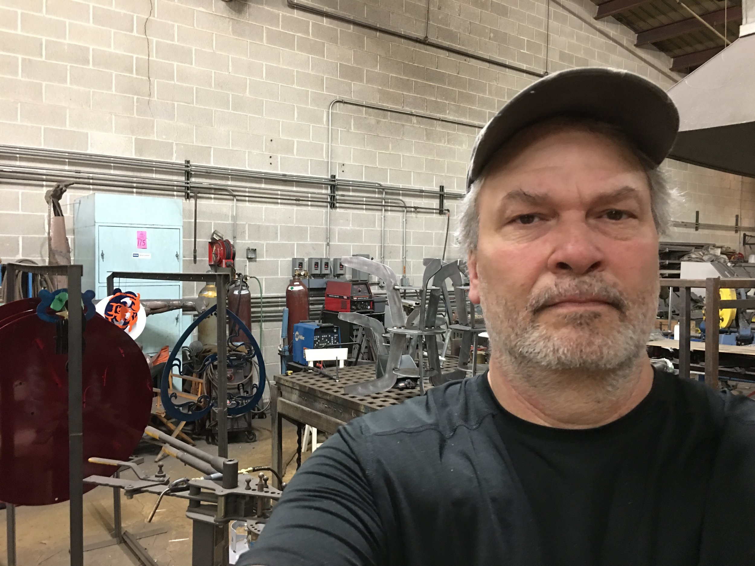 A man with gray hair and beard wearing a black shirt and gray cap taking a selfie in an industrial workshop or manufacturing facility with various machinery, metal parts, and equipment in the background.