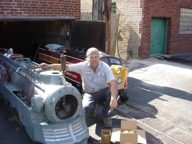 A worker crouching next to a robotic vehicle with a large metal wheel, on a city street with brick buildings, a utility pole, and a cardboard box nearby.